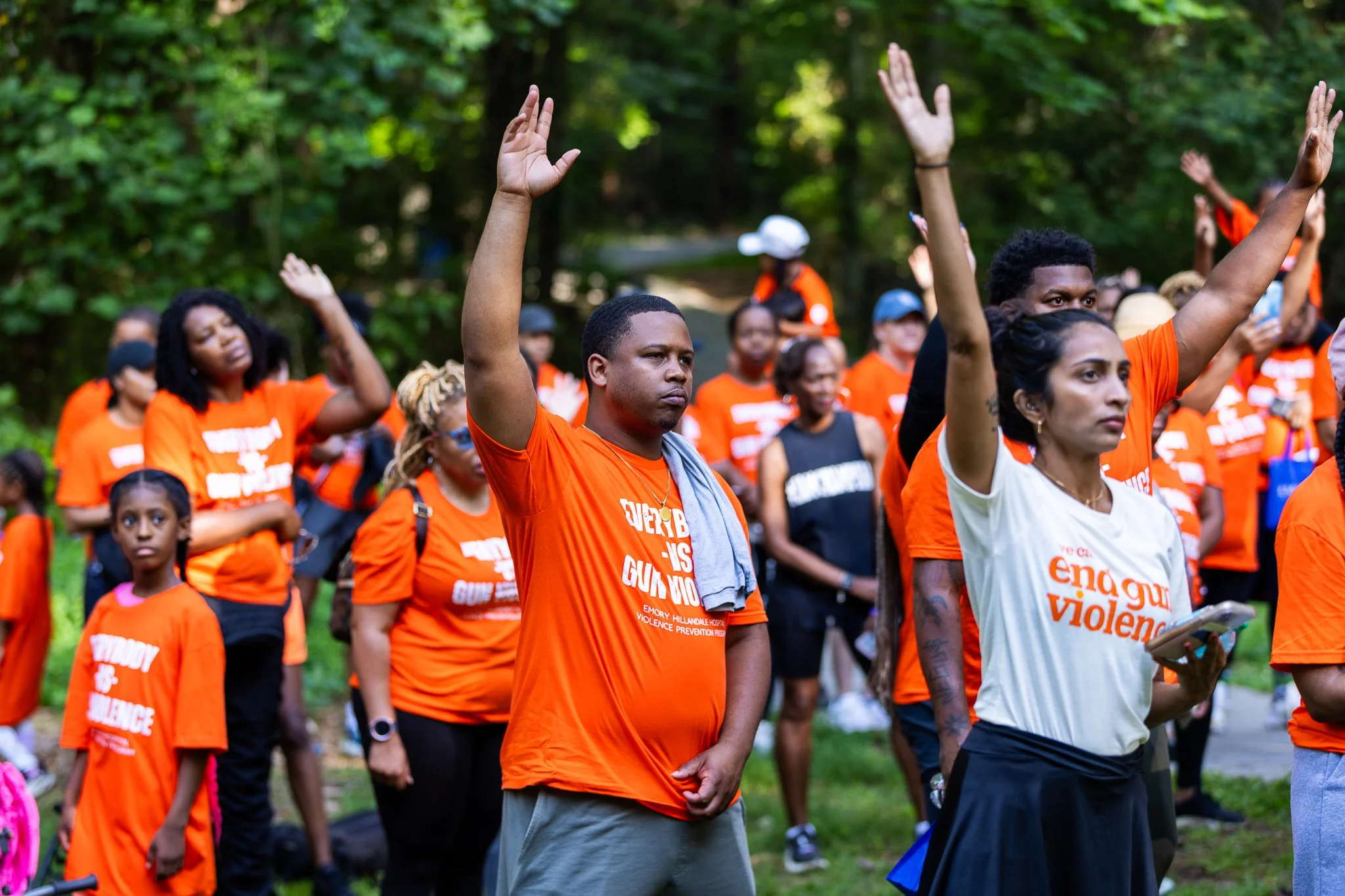 A group of protesters wearing orange shirts with a message about ending violence, standing outdoors in a wooded area, raising their hands in a gesture of protest.