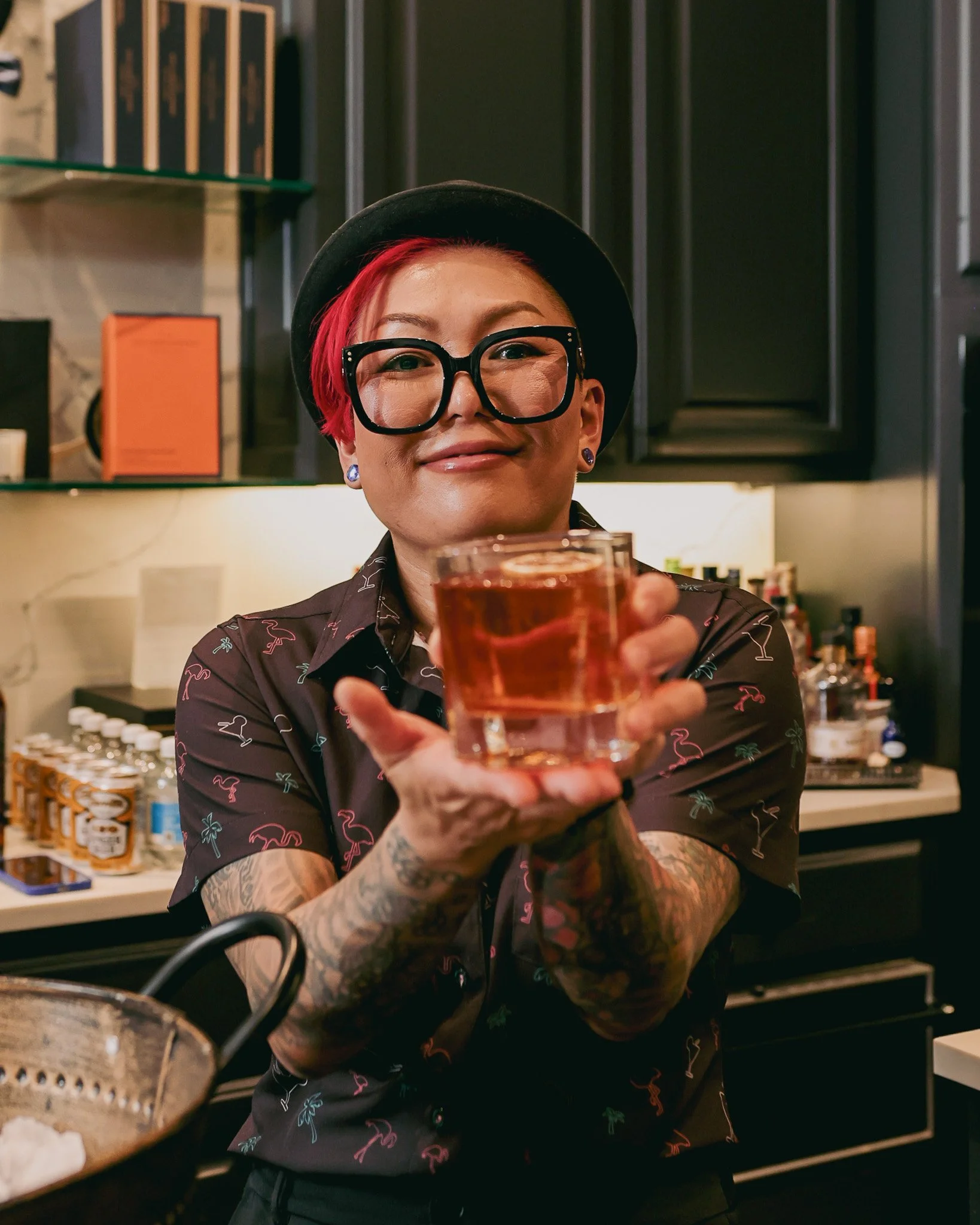 Person with pink hair, glasses, and tattoos holding a glass of drink, smiling at the camera in a bar or restaurant setting.