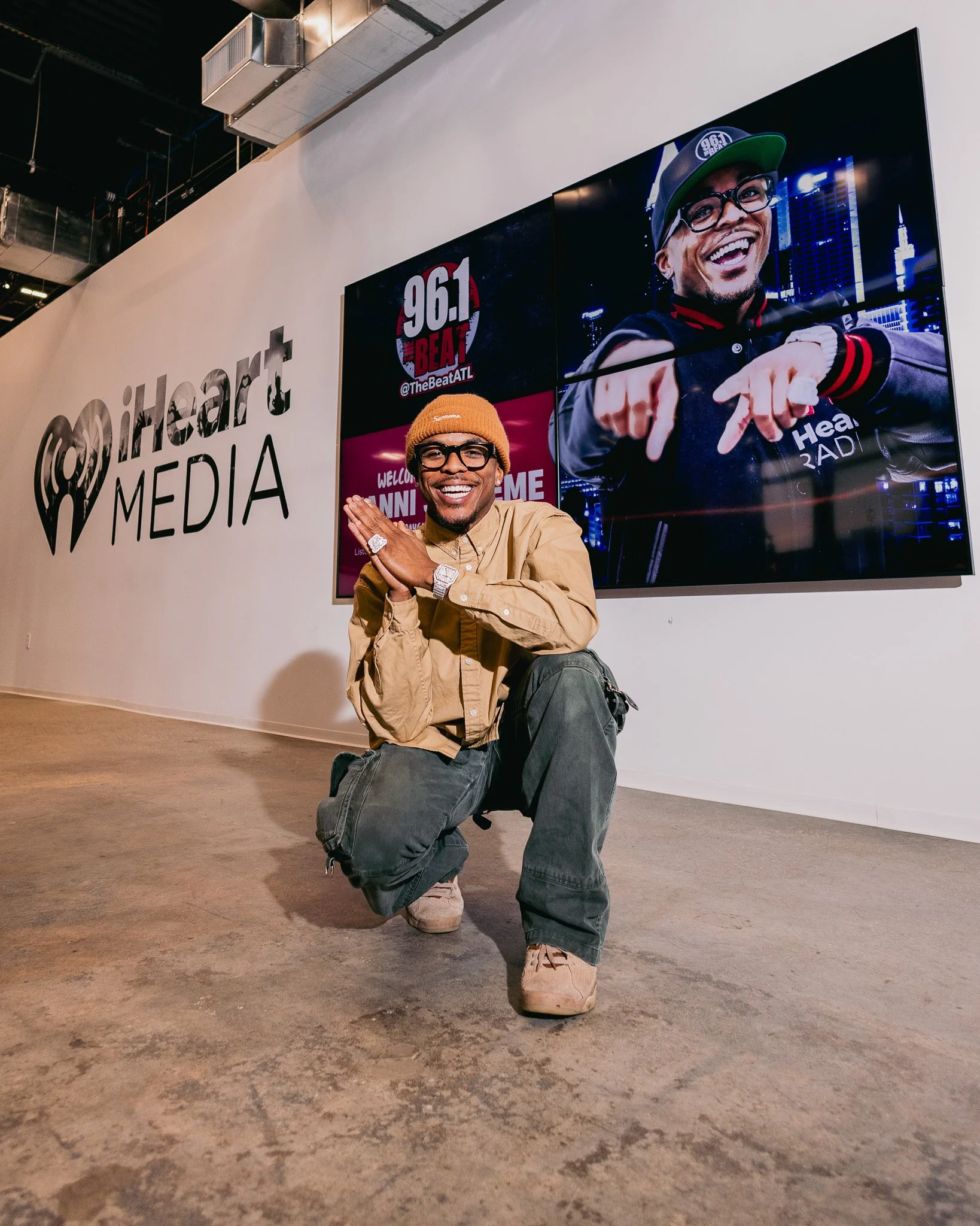 A man kneeling on one knee inside a media office or studio, smiling and posing. He is wearing glasses, a tan jacket, a yellow beanie, and sneakers. Behind him is a large screen displaying a smiling man in a jacket and cap, with city buildings in the 
