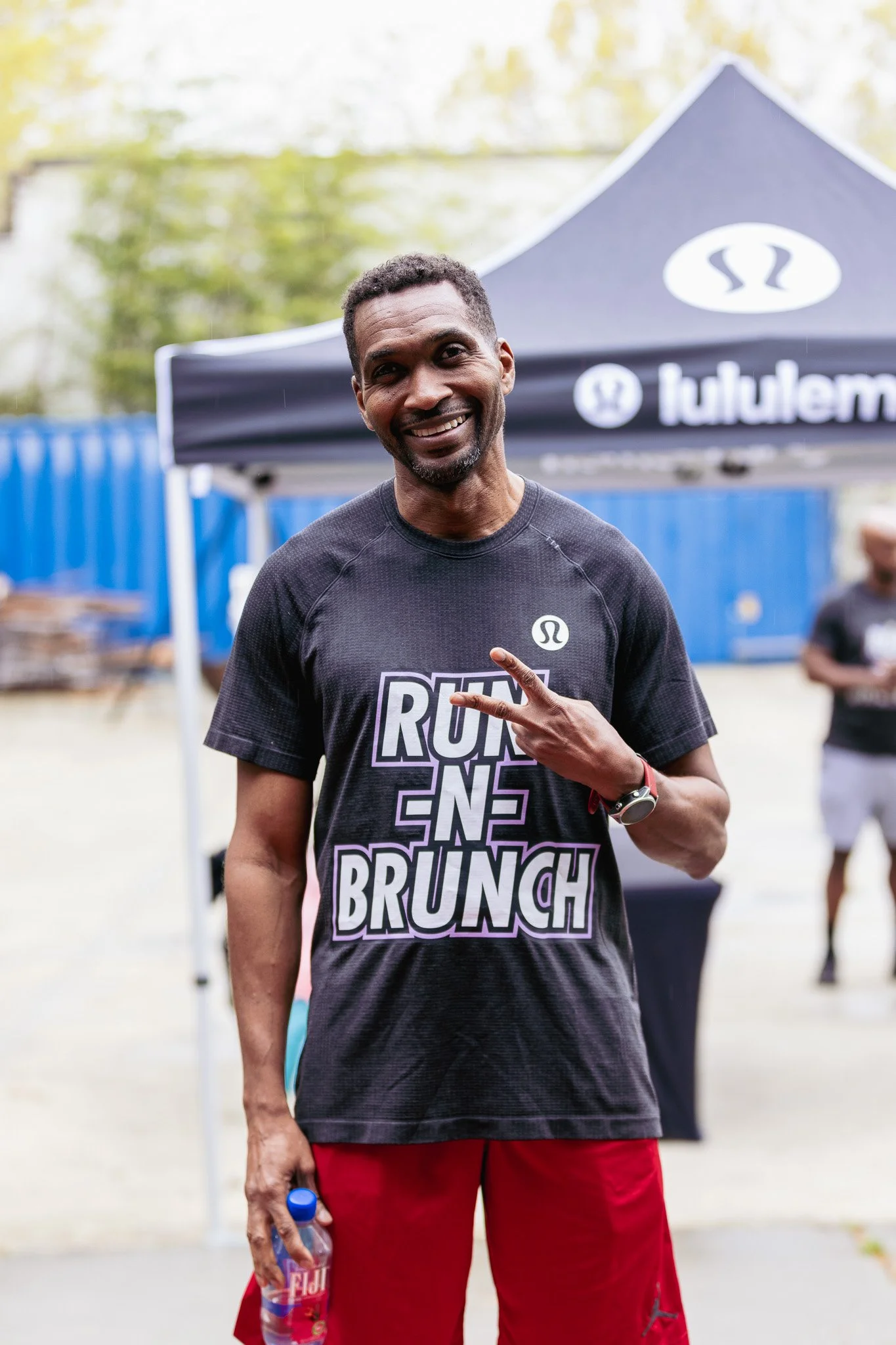Smiling man wearing black T-shirt with words 'RUN ENDS BRUNCH' and red shorts, holding a water bottle, standing outdoors near a blue tent with a logo.