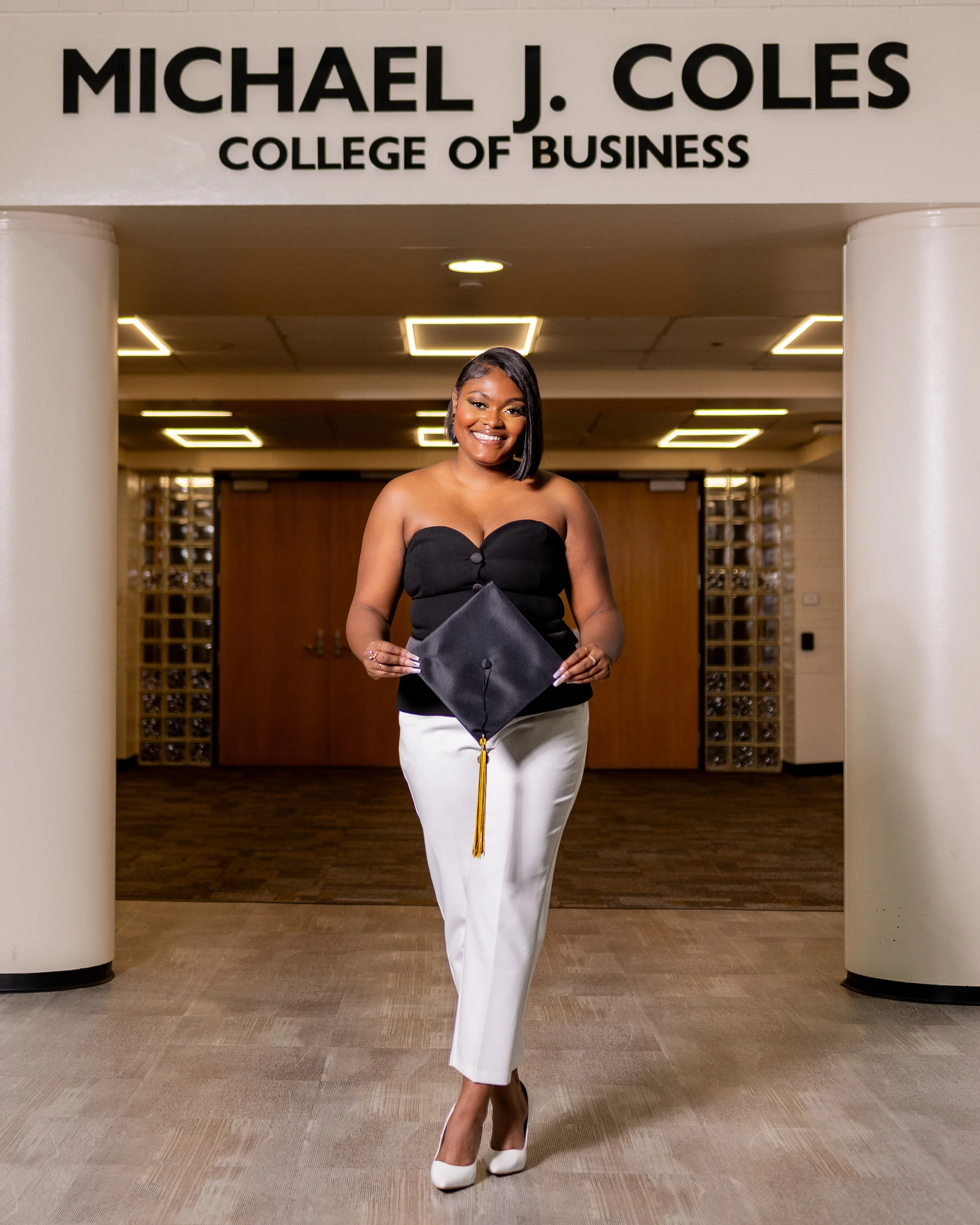 A young woman with dark hair styled in a bob, smiling, dressed in a strapless black top, cream-colored pants, and white heels, holding a doctoral cap with a gold tassel, standing in front of a sign for Michael J. Coles College of Business in an indoo