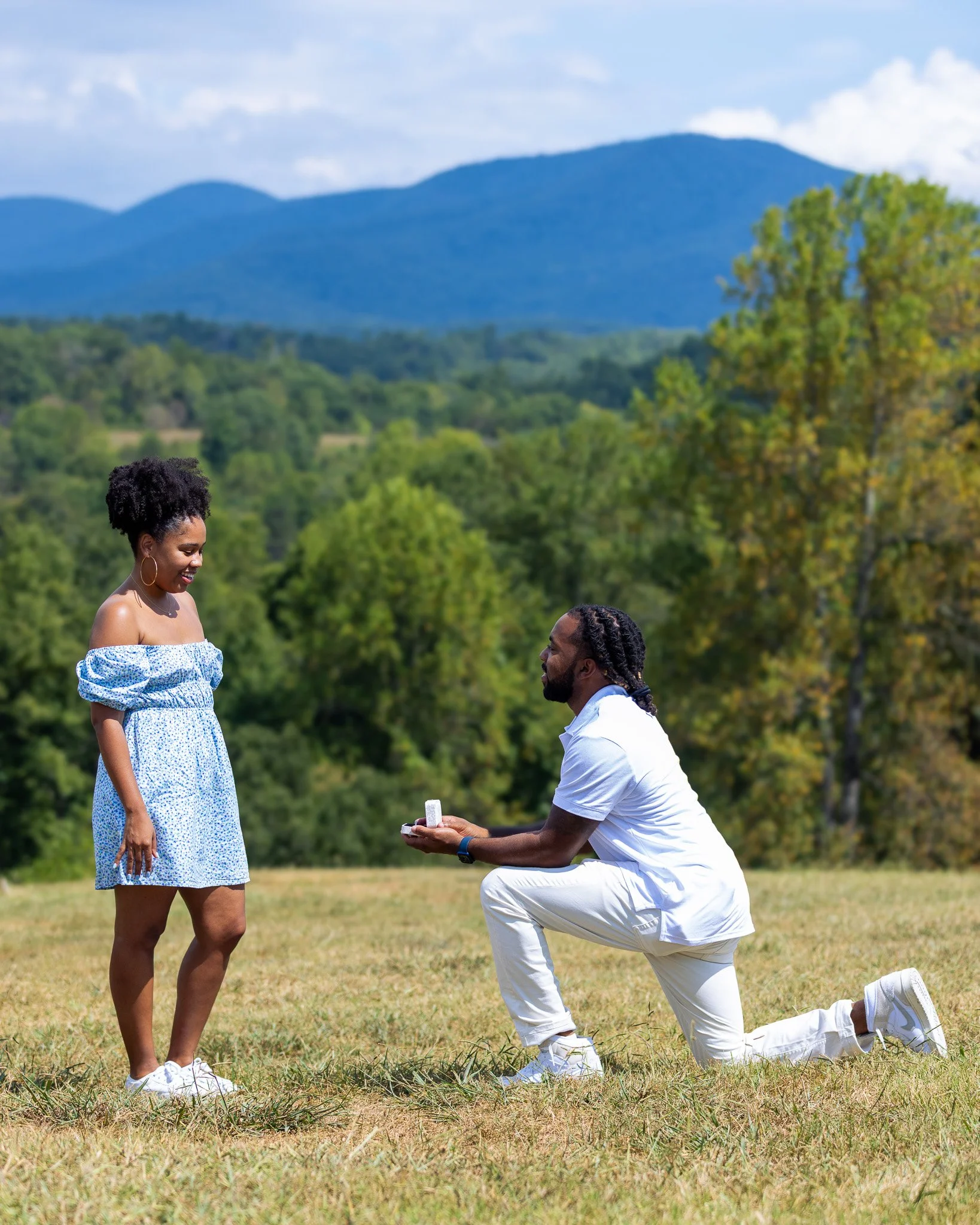 A man kneeling on one knee proposing with a ring to a woman standing outdoors in a park with trees and mountains in the background