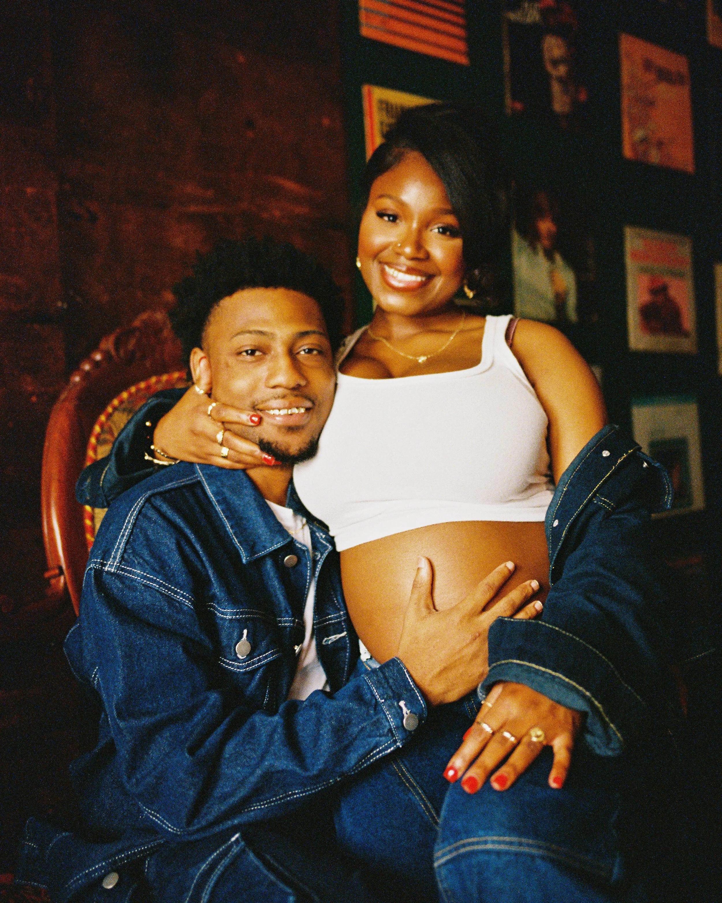 A smiling woman with earrings and a necklace sitting next to a man with short hair and facial hair, both wearing denim jackets, with the woman's hand on her pregnant belly, in a room with framed pictures on the wall.