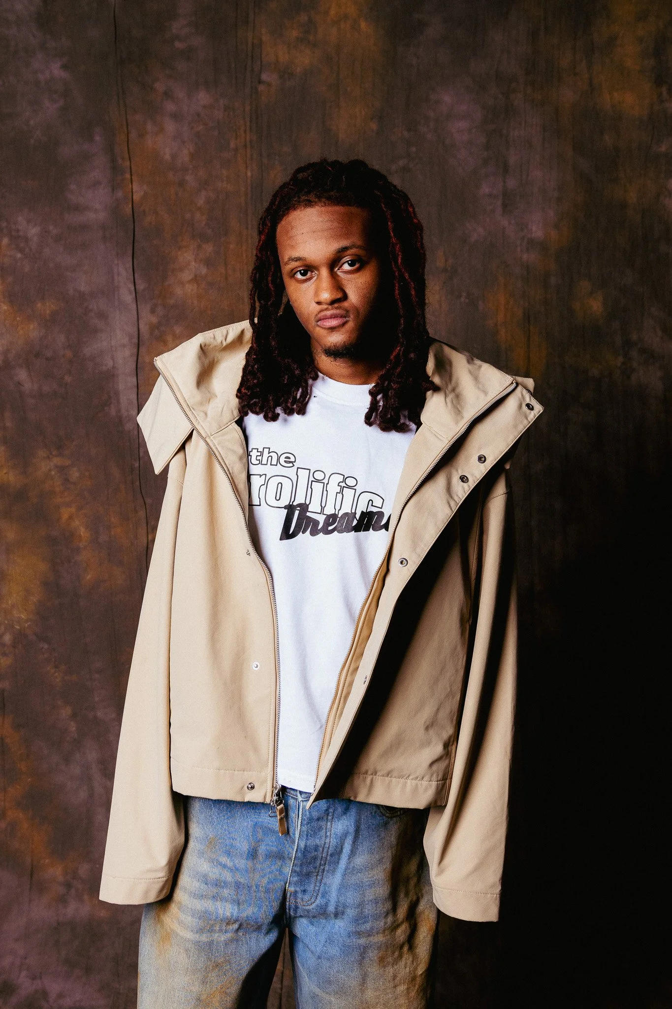 A young man with long dreadlocks wearing a beige trench coat over a white t-shirt with text, and light blue jeans, standing in front of a dark, textured background.