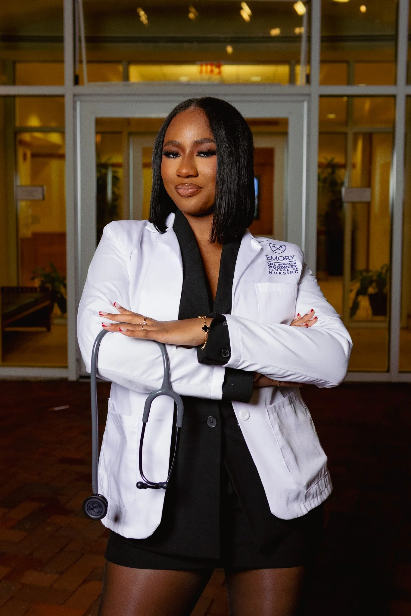 A woman wearing a white medical coat with EMORY University and Nell Hodgson Woodruff School of Nursing embroidery, posing with arms crossed and a stethoscope around her neck, standing in a hospital or clinic corridor.