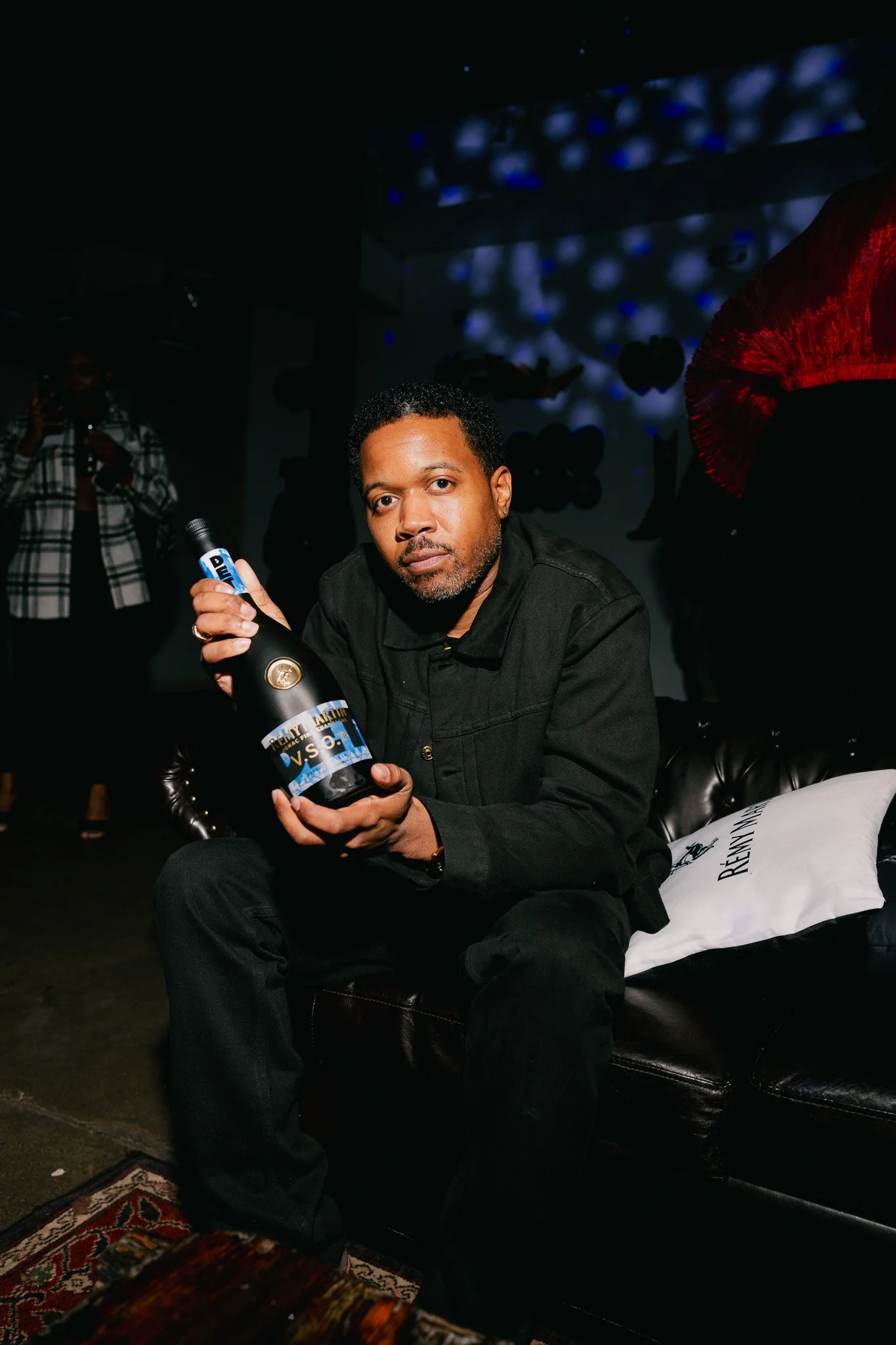 A man sitting on a black leather couch holding a bottle of Veuve Clicquot champagne at a party or celebration. There are balloons and decorative elements in the background, with low lighting and colorful projected lights on the ceiling.