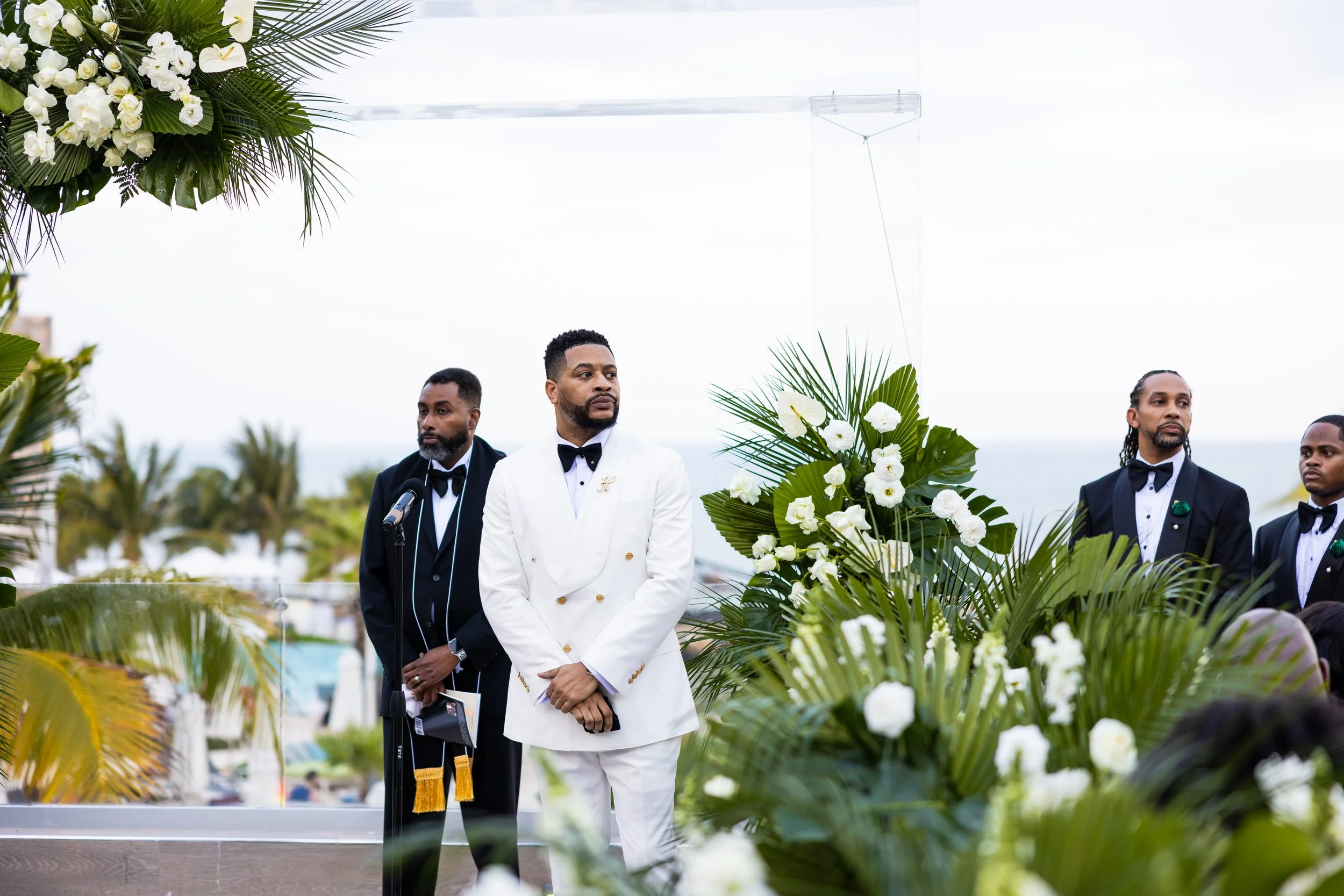 A groom in a white tuxedo and a group of groomsmen in black tuxedos standing outdoors during a wedding ceremony, with white floral arrangements and greenery.