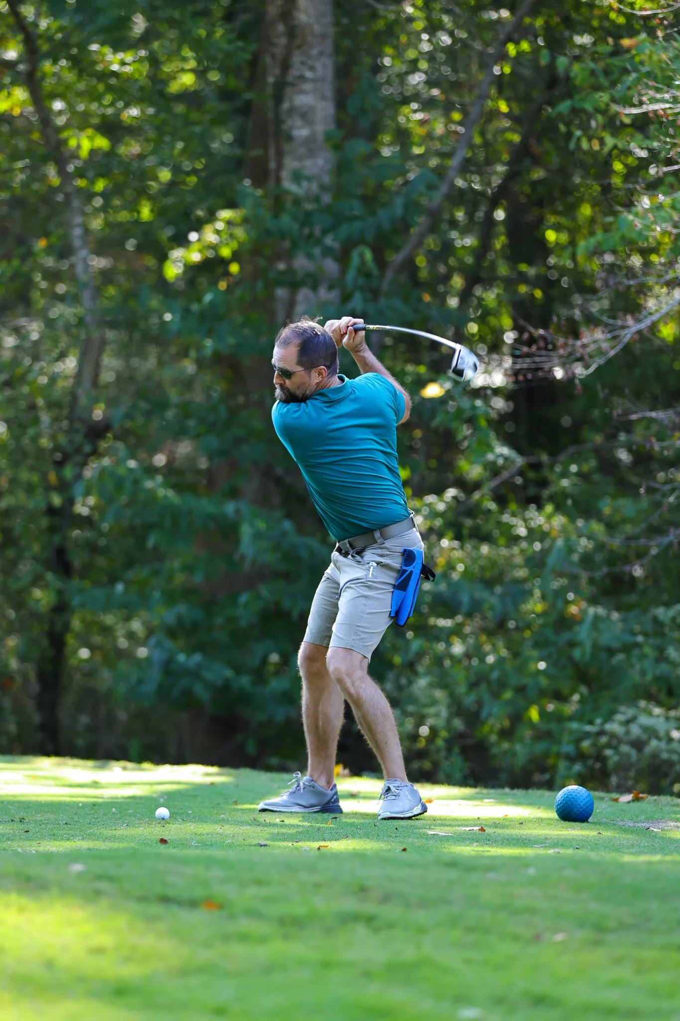A man playing golf on a lush green golf course, preparing to hit a golf ball with a driver club. The man is wearing sunglasses, a blue shirt, khaki shorts, and gray golf shoes. There is a blue golf tee and a blue golf ball on the grass, with trees an