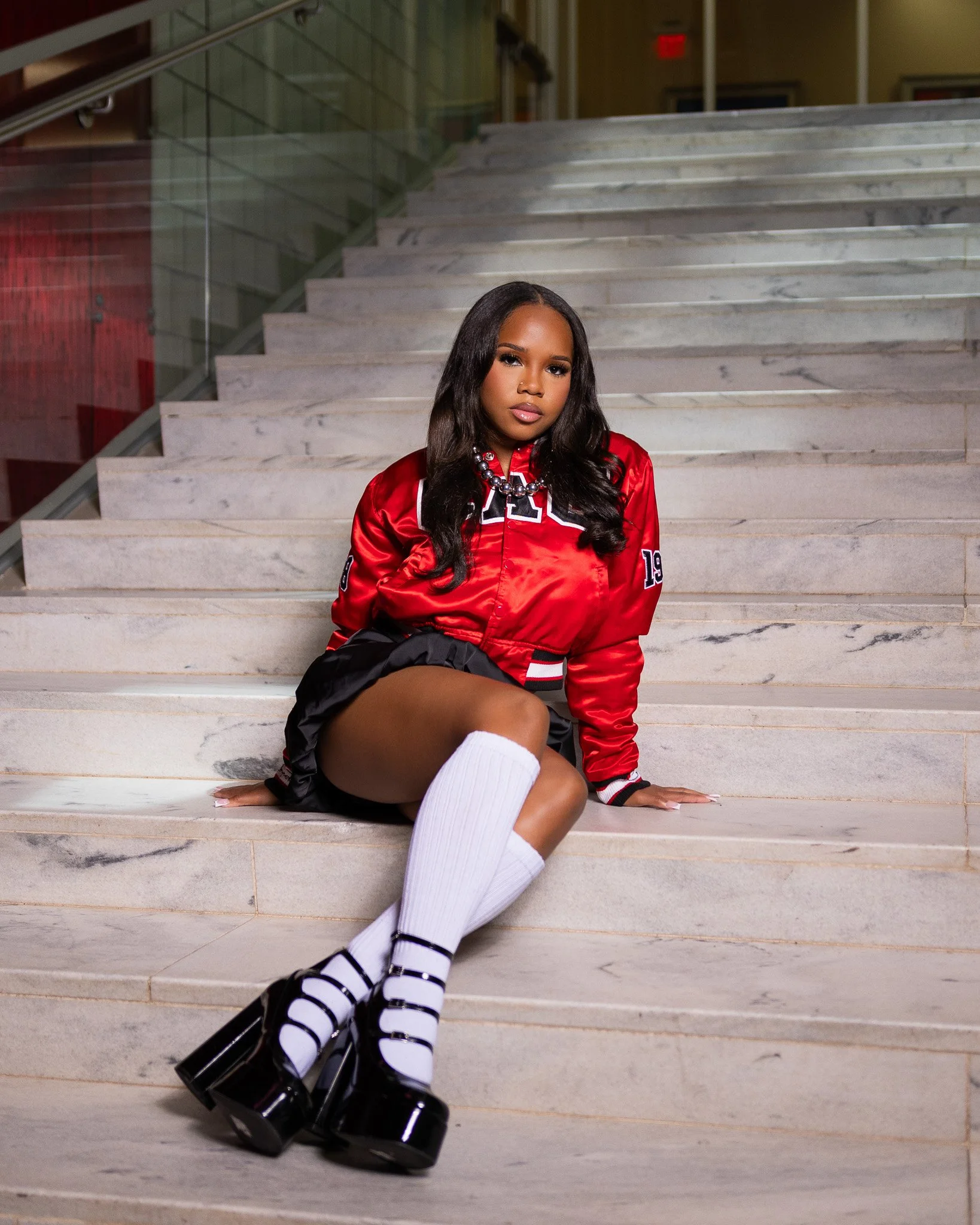 A young woman wearing a red sporty jacket, black shorts, white knee-high socks, and black shoes with white straps, sitting on marble stairs in an indoor setting with a glass railing and a yellow wall in the background.