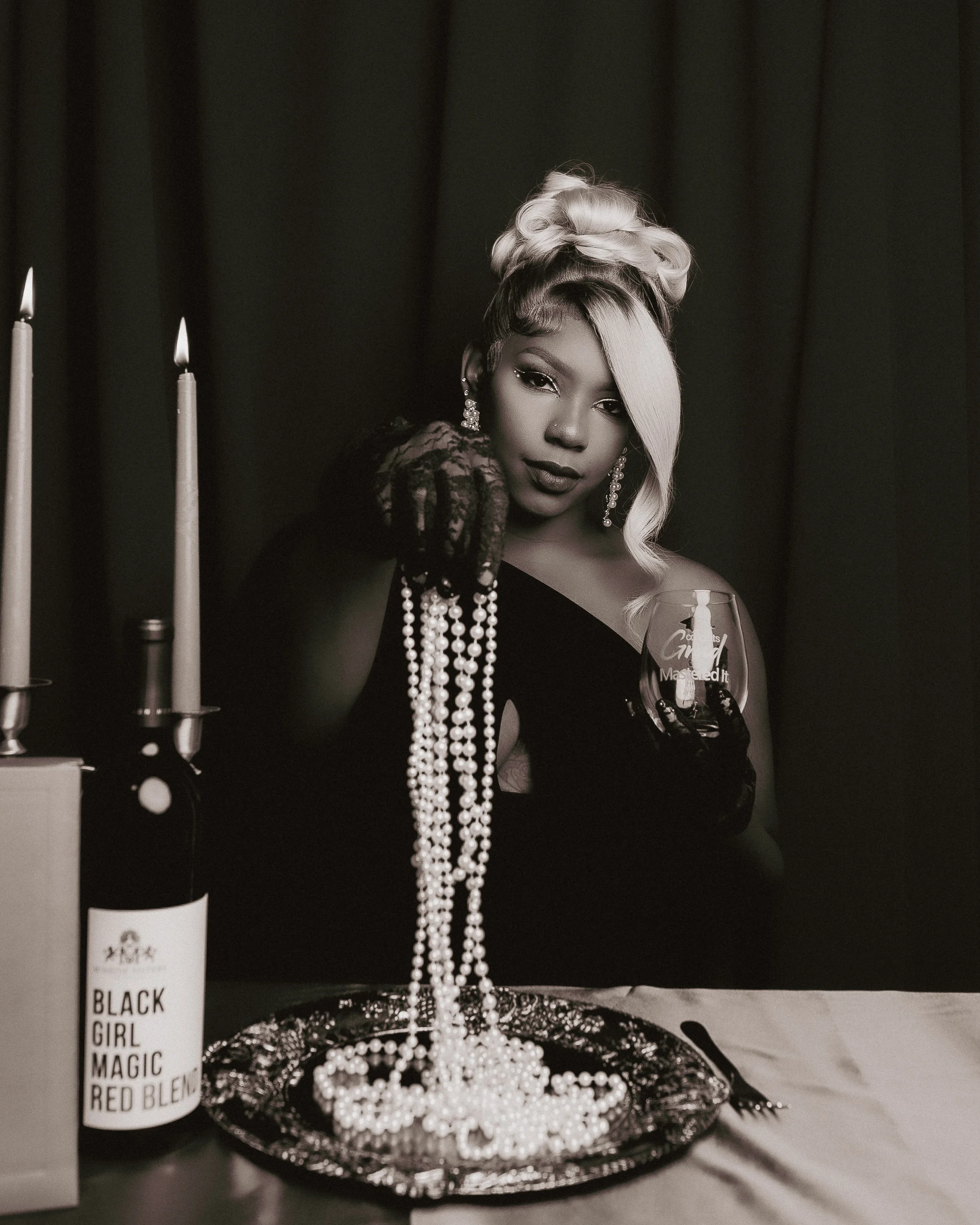 Black and white photo of a woman with elegant hair and jewelry, holding a glass of wine, surrounded by pearls on a decorative tray, with a bottle of wine labeled 'BLACK GIRL MAGIC' on the side.