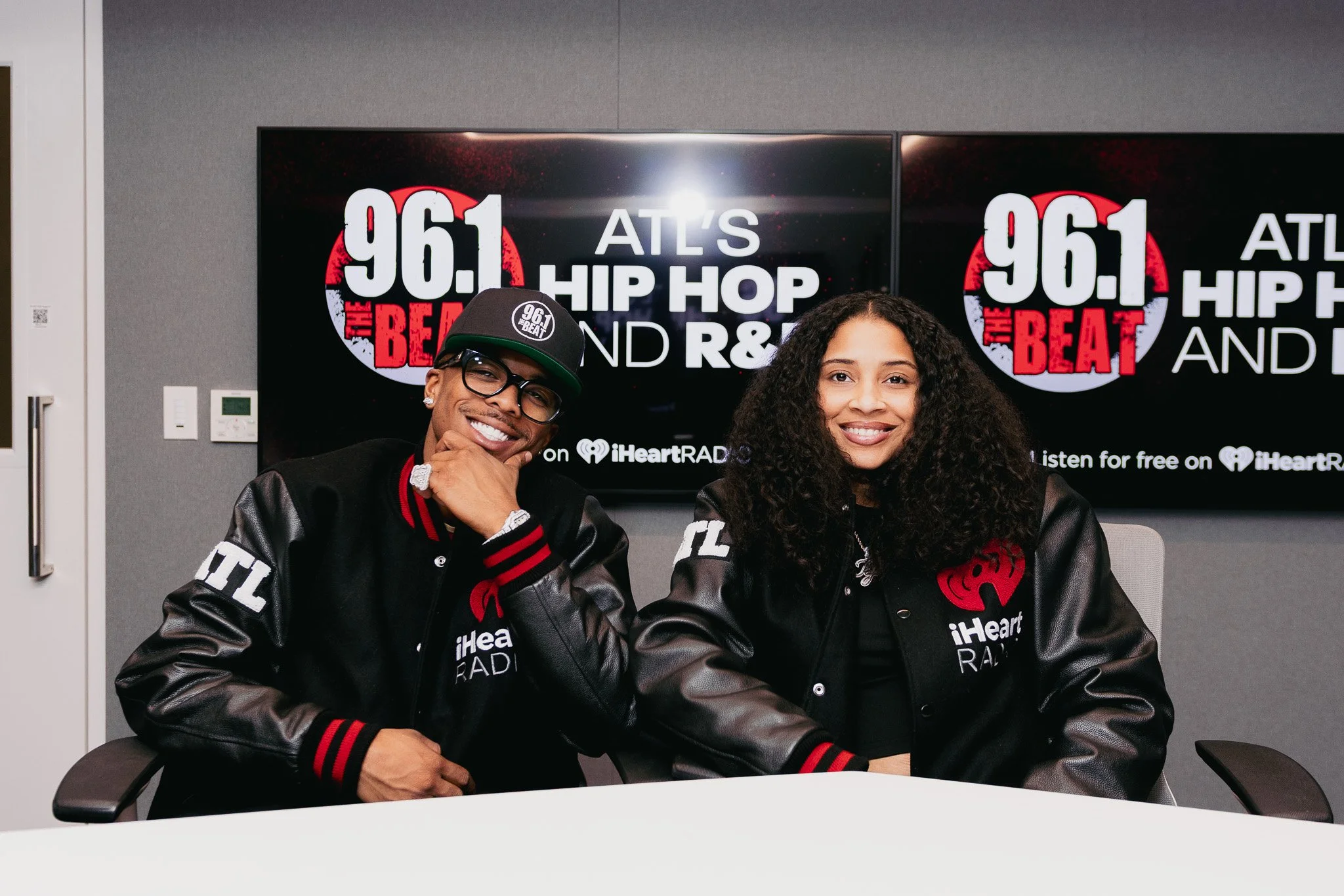 Two people sitting at a table, smiling in front of a large screen with radio station information. Both are wearing black jackets with logos for iHeartRadio and 96.1 The Beat. The man wears glasses and a black cap with the station's logo, and the woma