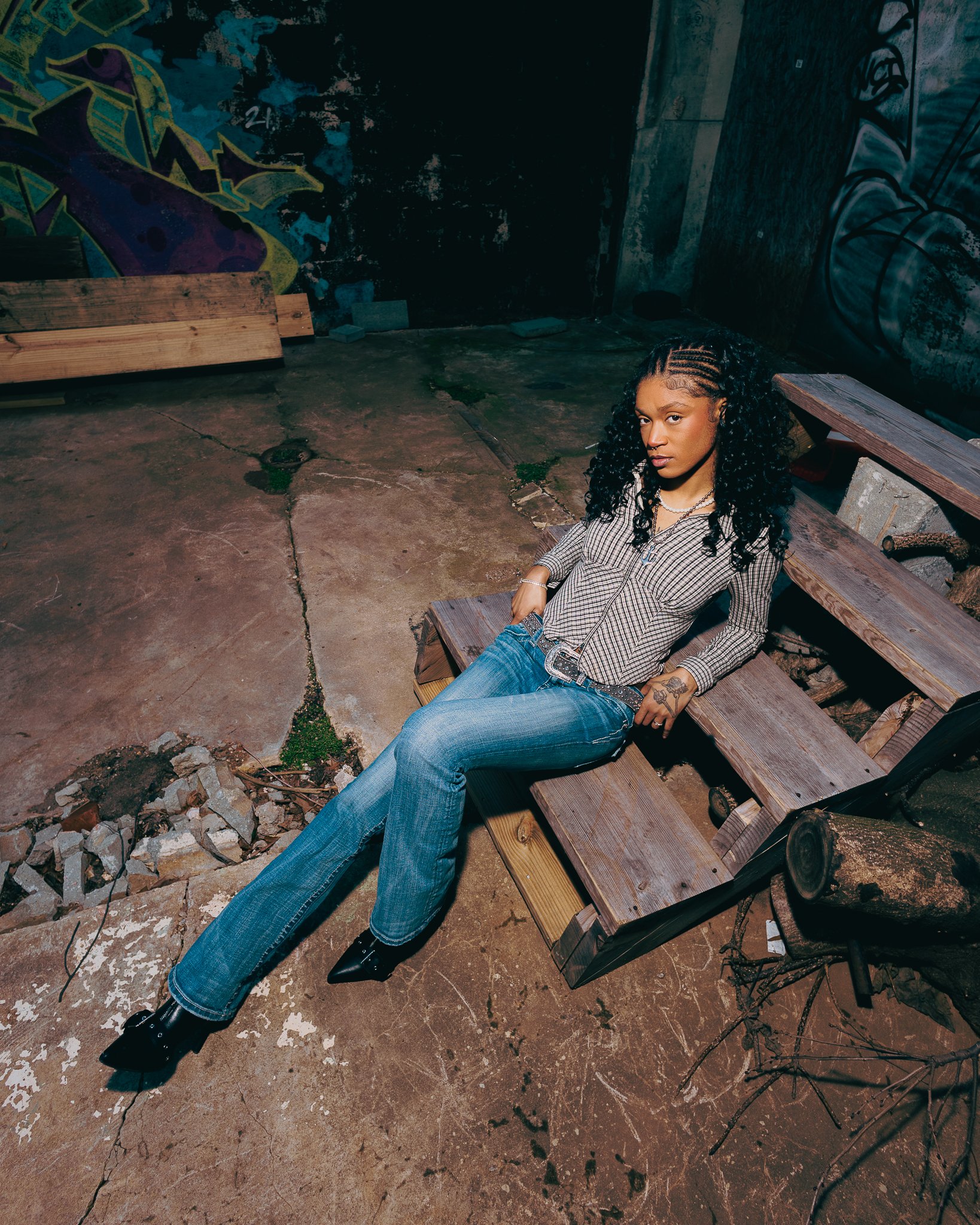 A young woman with curly hair and a checked shirt sitting on a wooden bench in an urban outdoor setting, with graffiti artwork on a wall behind her and a concrete floor.