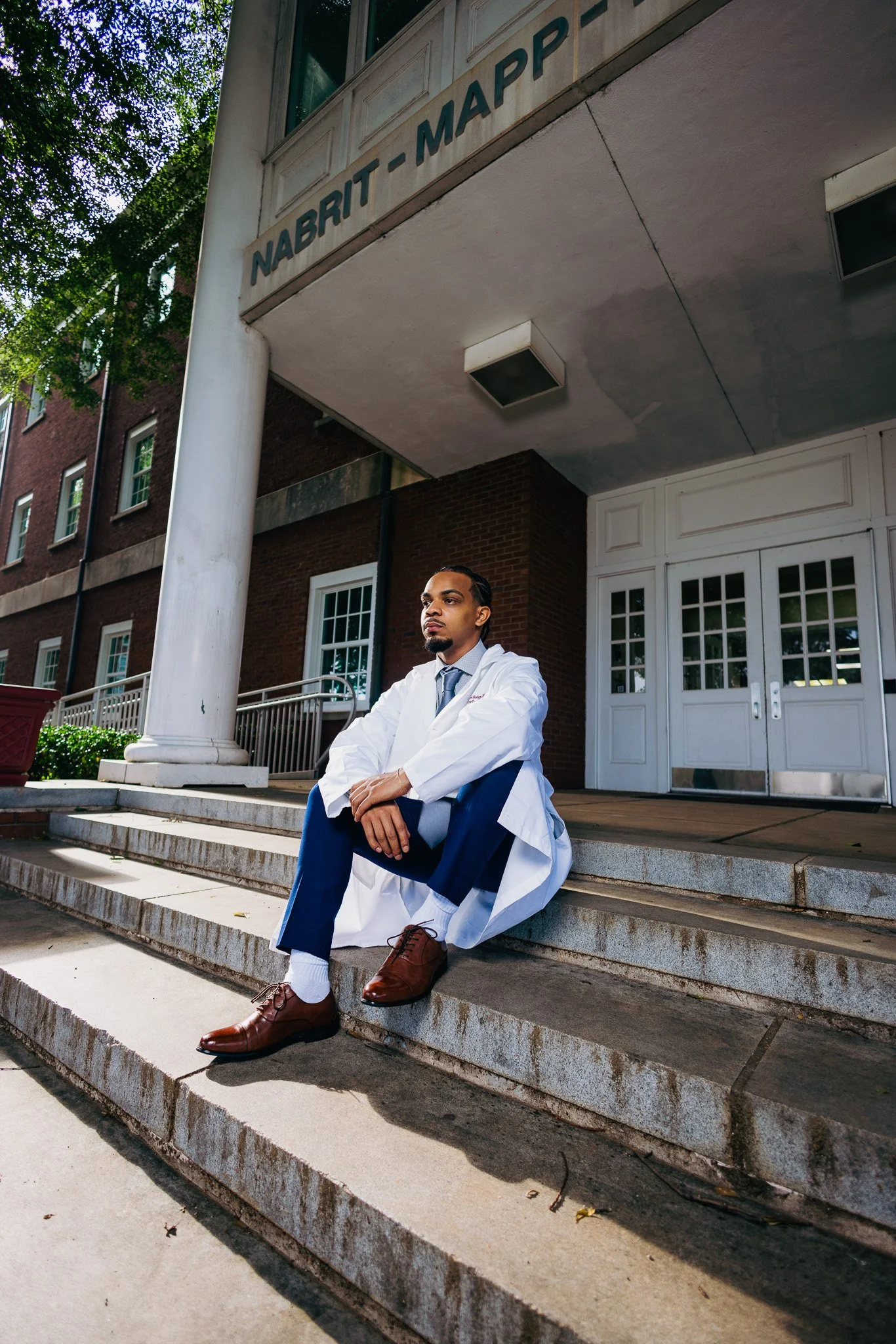 A man in a doctor's coat sitting on the front steps of a building with a sign that reads "NABRIT - MAPPT." The building has brick walls, white doors, and windows, with trees and a blue sky visible in the background.