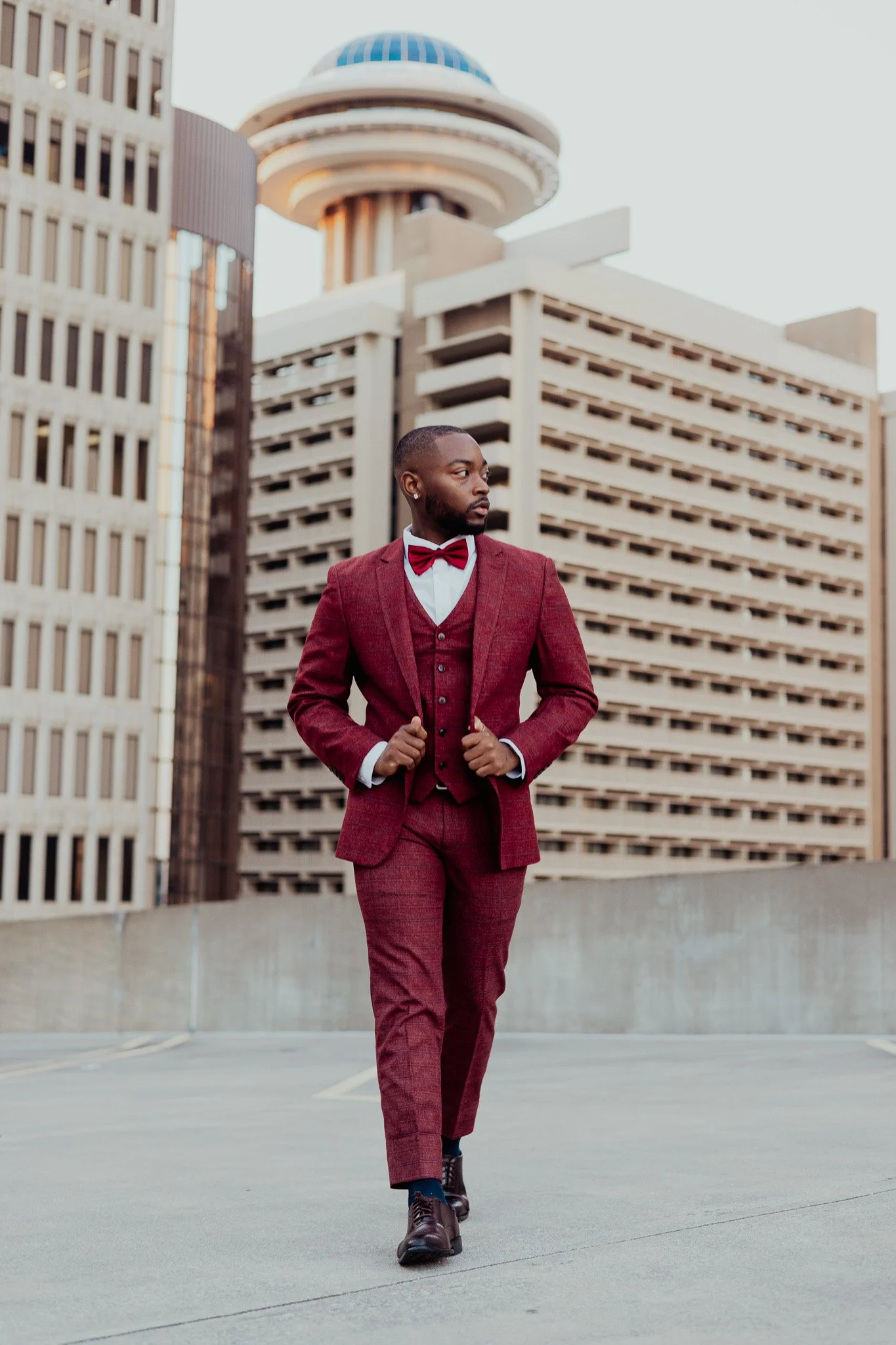 A man wearing a red suit, white shirt, and red bow tie walking on a rooftop with high-rise buildings in the background.