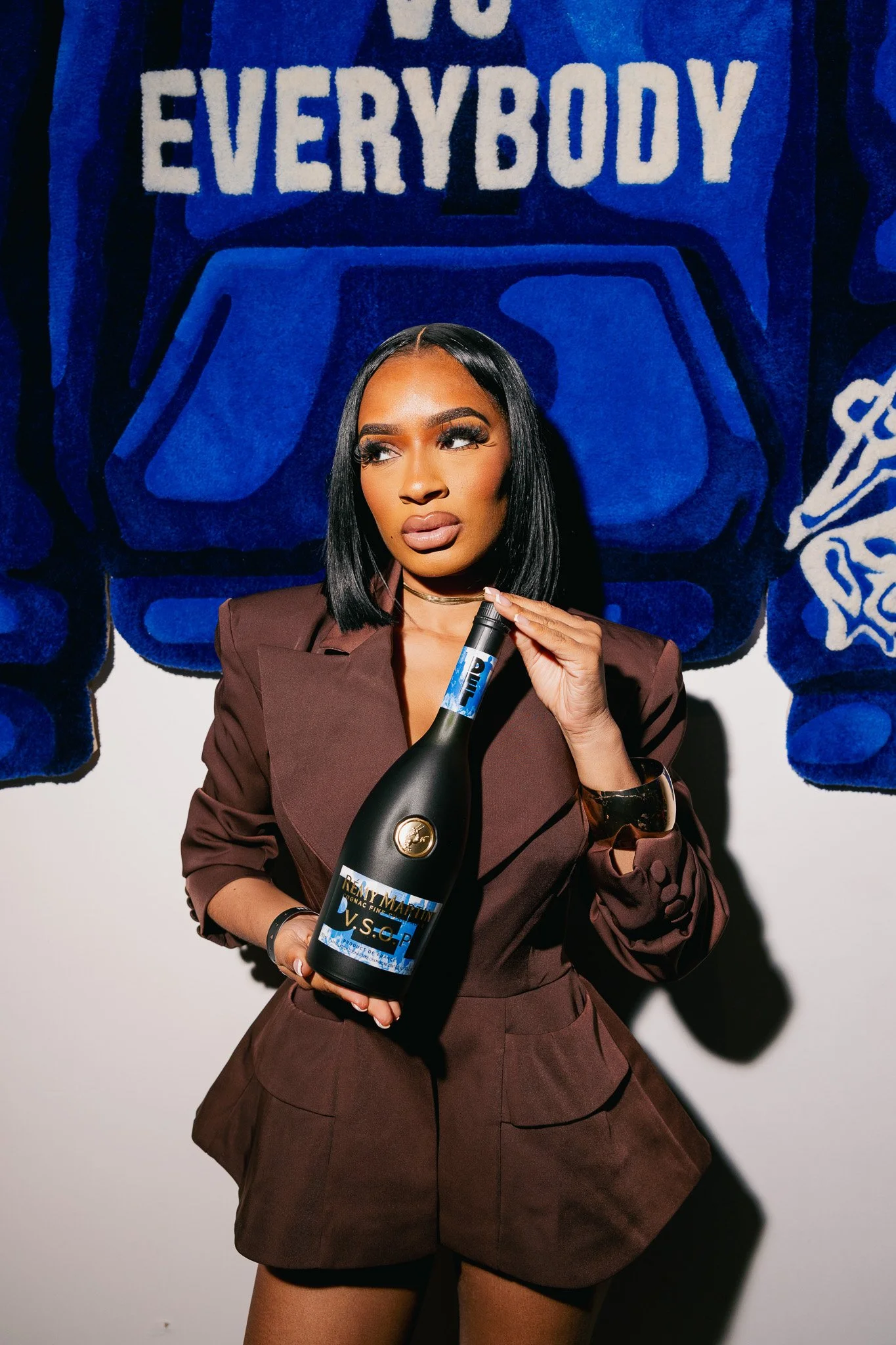 A woman with dark hair and makeup holding a black bottle of Remy Martin VSOP cognac, standing in front of a blue and black textured background with the words 'Everybody' visible at the top.