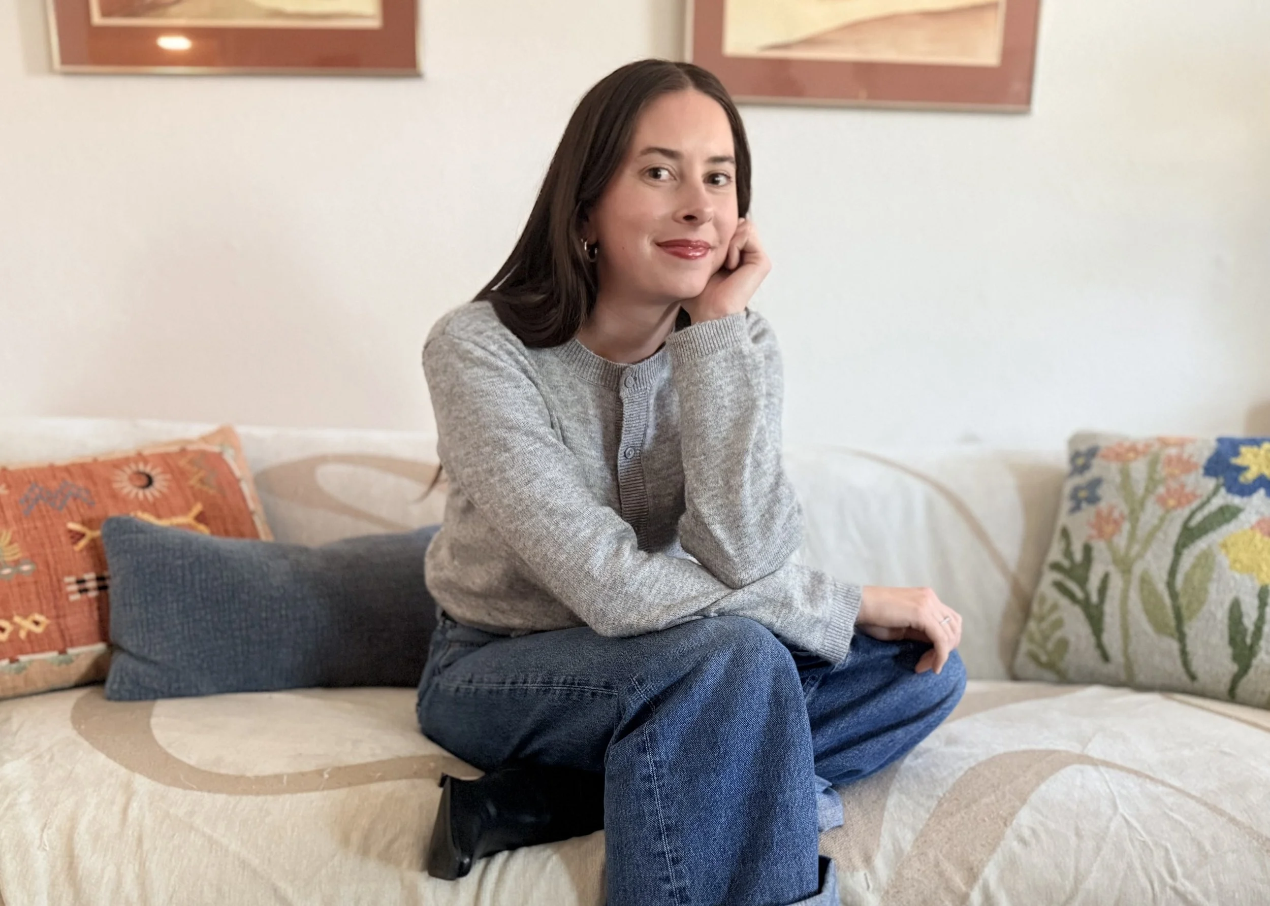 A woman with long dark hair, sitting on a couch, resting her head on her hand, smiling, wearing a gray sweater and jeans, surrounded by colorful pillows in a living room.