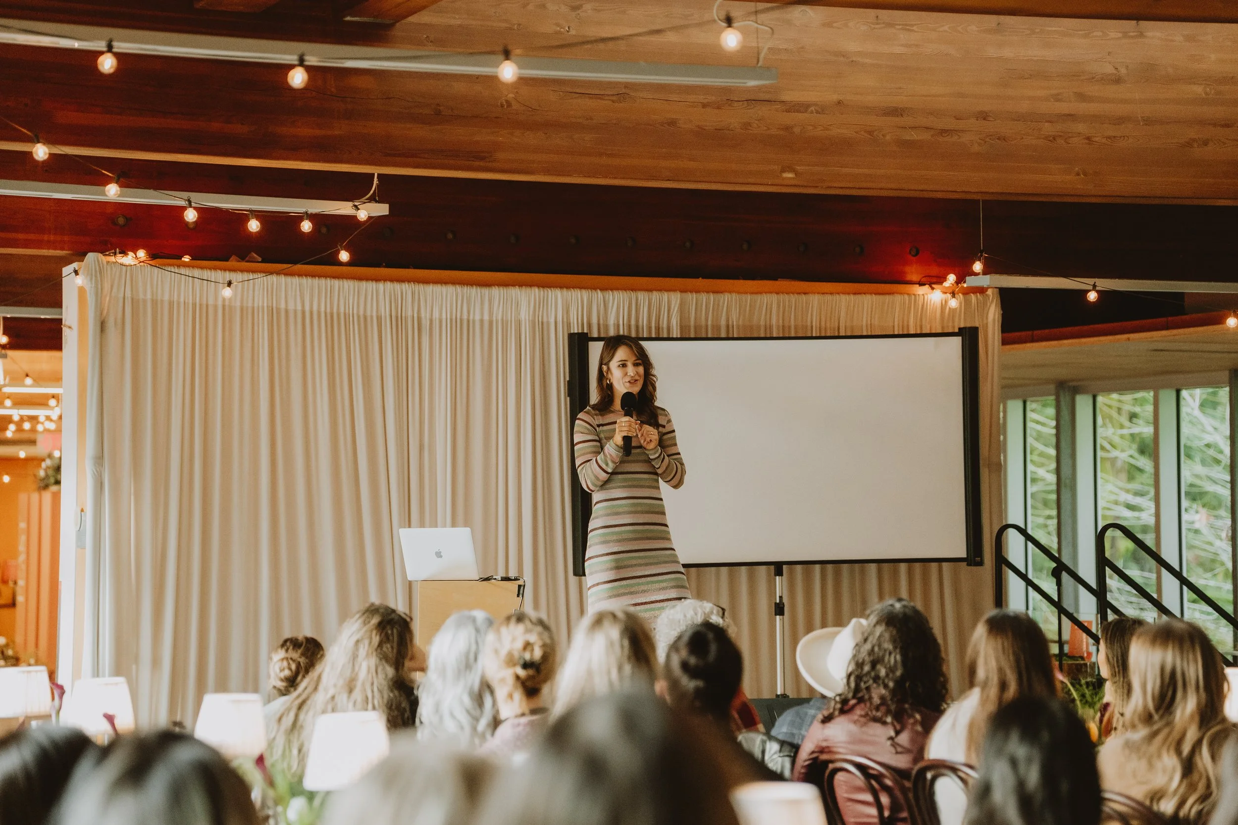 A woman in a striped dress appears to be speaking or giving a presentation in front of a seated audience in a room with wood-paneled ceilings and string lights.