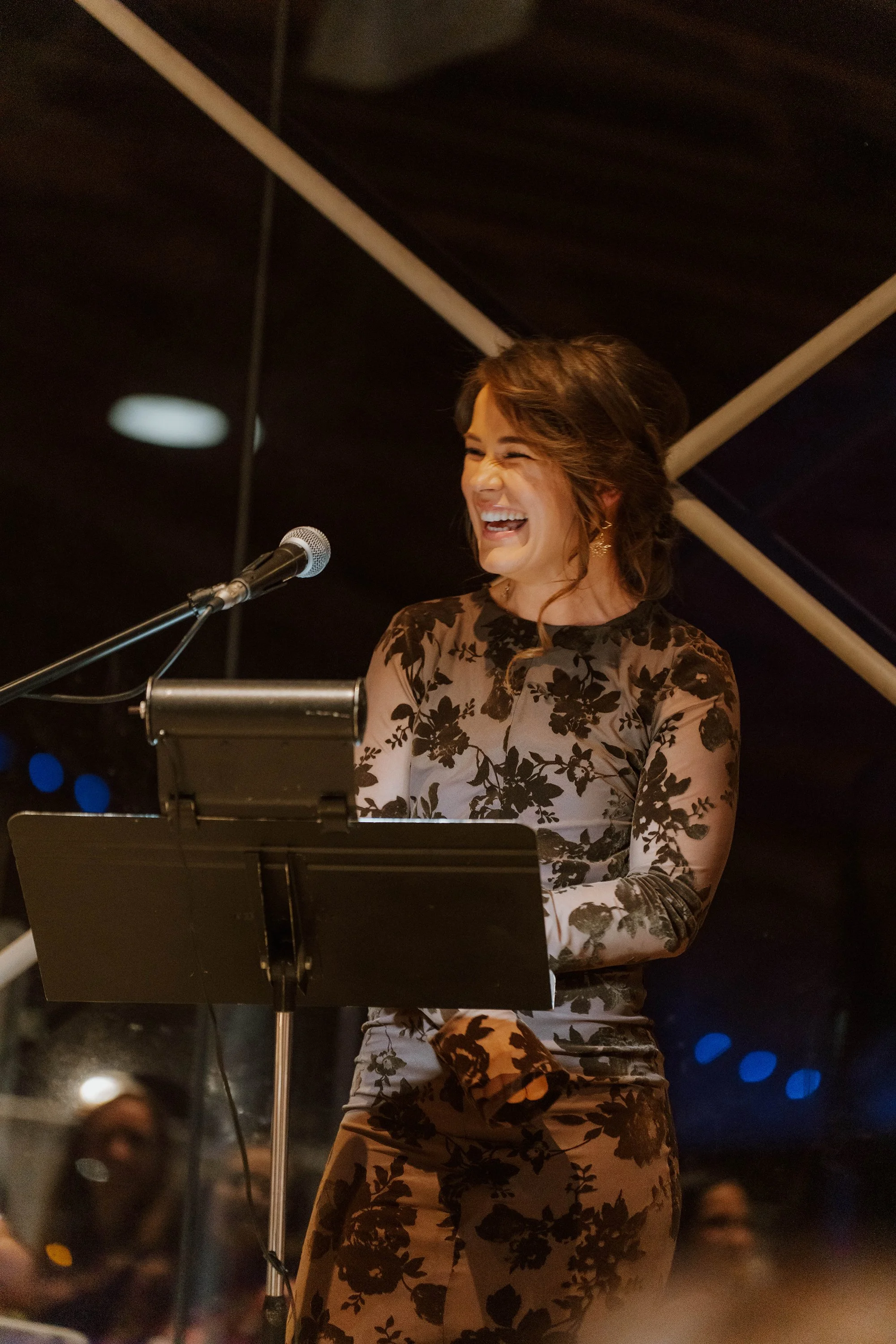 A woman wearing a floral-patterned dress is singing or speaking into a microphone at a podium indoors, with a blurred audience visible in the background.