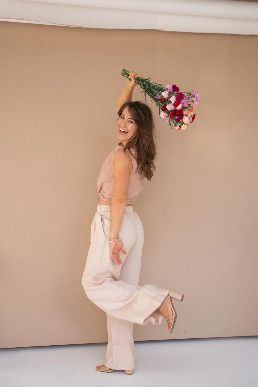 A woman standing against a plain beige wall, smiling and holding a bouquet of pink and red flowers over her head, wearing light-colored, loose pants and a sleeveless top.