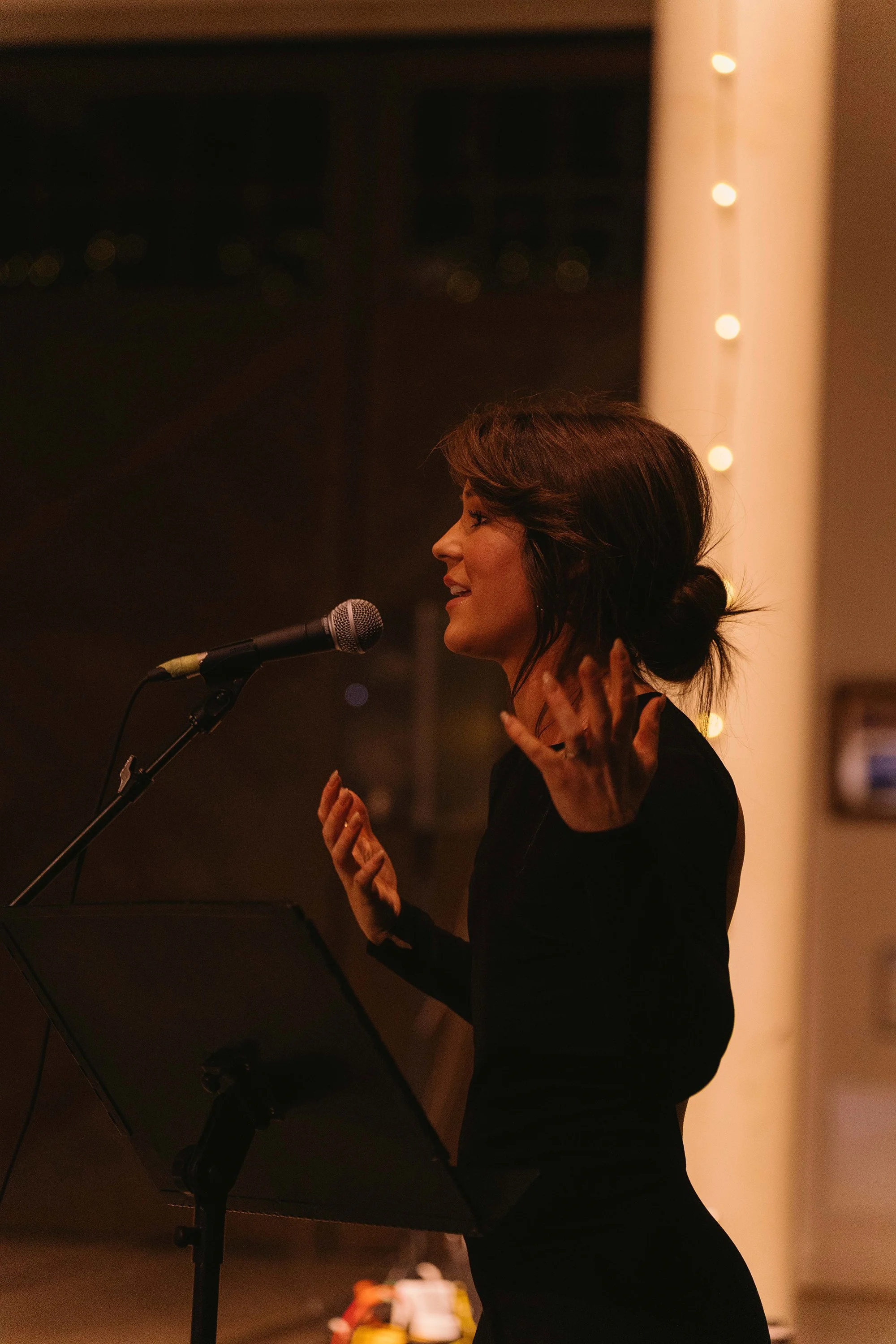 A woman with shoulder-length brown hair in a bun, wearing a black outfit, singing passionately into a microphone with eyes closed, inside a warmly lit room decorated with string lights.