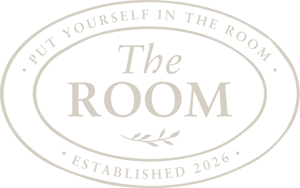 A black oval sign with light gray text that reads "Put Yourself in the Room" at the top and "Established 2026" at the bottom. In the center, it says "The Room" with a small decorative branch beneath.