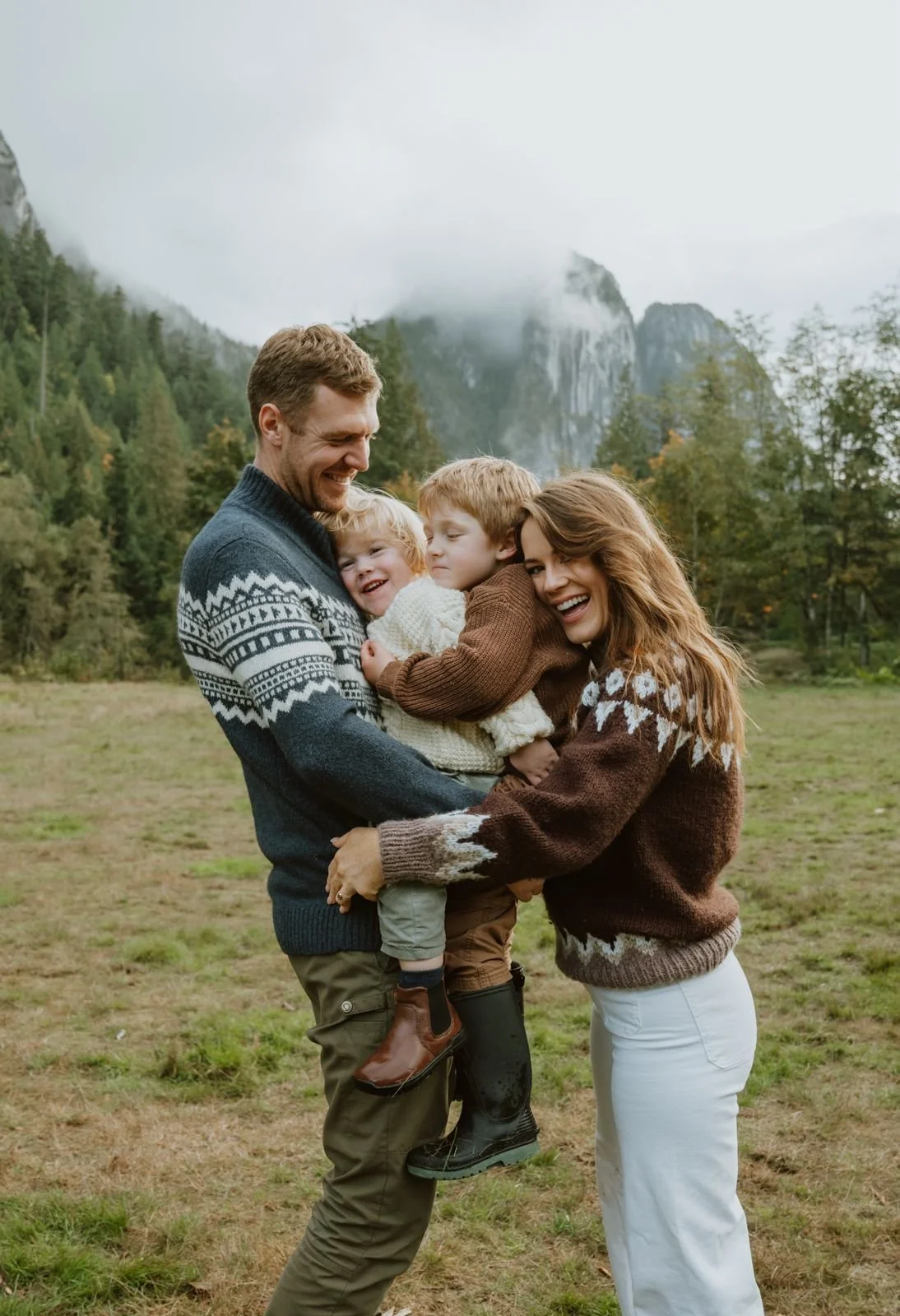 Family of four enjoying outdoors in a grassy field with mountains and trees in the background. The father and mother are holding two children, all smiling and dressed in cozy sweaters.