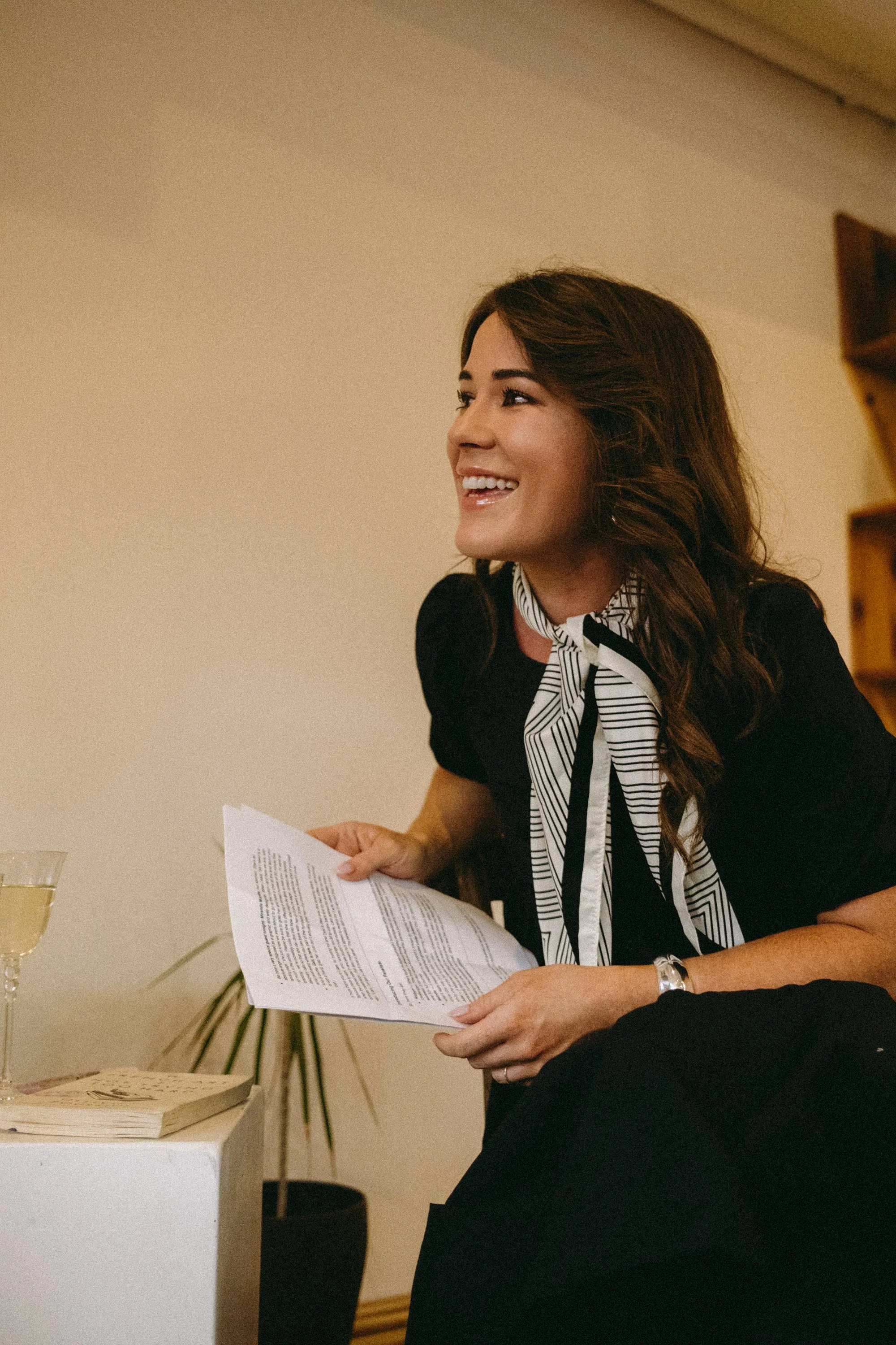 A woman with long dark hair, smiling, wearing a black outfit and a black and white striped tie, holding a paper, sitting at a table with a glass of wine, books, and plants nearby.