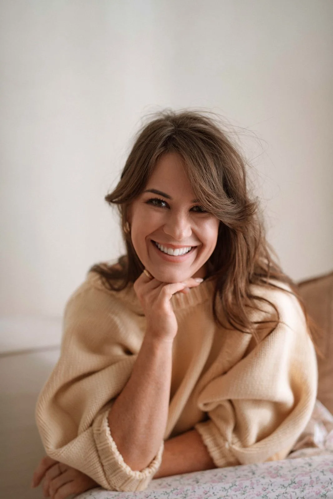 A woman with brown hair smiling, wearing a beige sweater, resting her chin on her hand, sitting indoors against a plain wall.