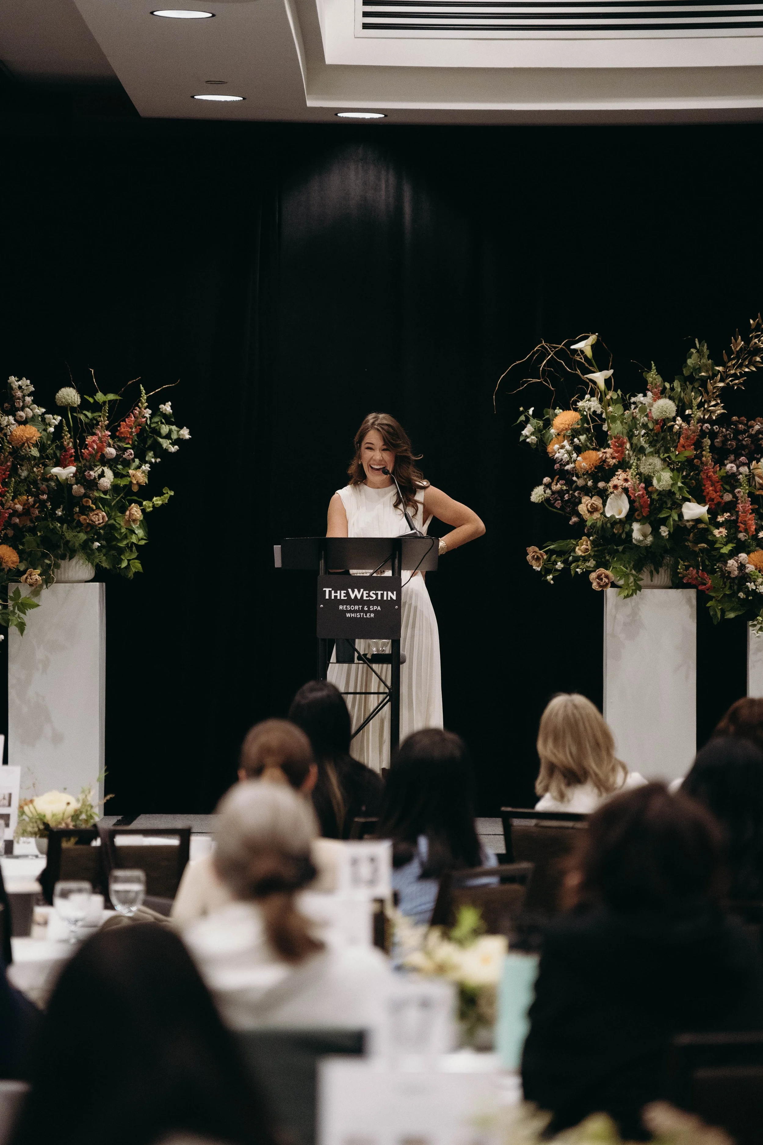 A woman speaking at a podium during an event at The Westin resort and spa in Whistler, with audience members seated at tables in front. There are large floral arrangements on either side of the stage, and the woman appears happy and engaged.