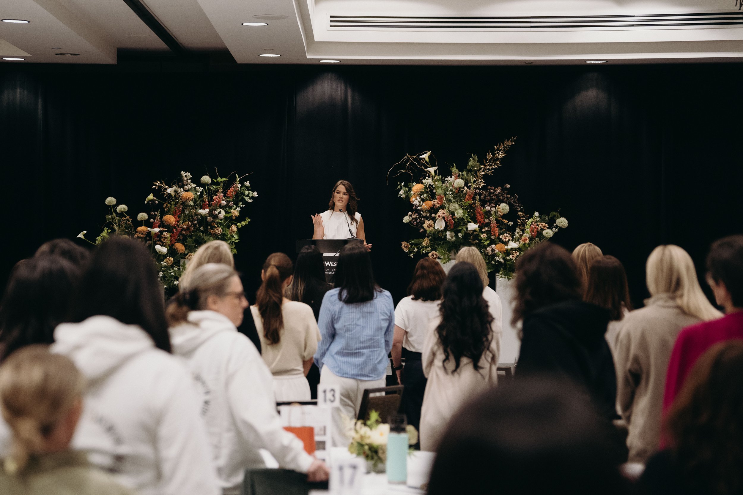 A woman giving a speech at a podium in front of a large audience in a conference room decorated with two tall floral arrangements.
