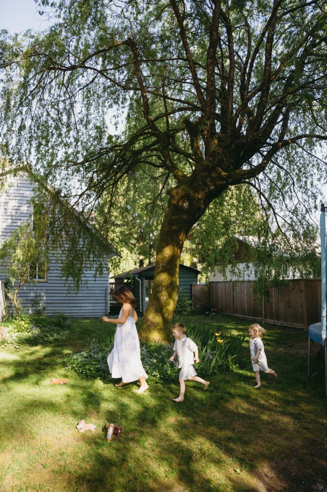 A woman and two children, all dressed in white, running and playing in a sunny backyard with a large tree, green grass, and a wooden fence.