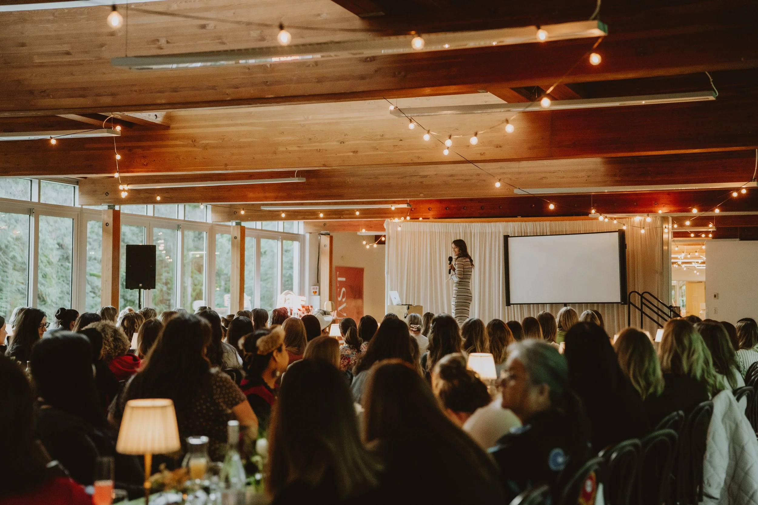 Interior of a conference room filled with an audience. A woman stands on stage holding a microphone near a large white screen. The room has wooden ceiling panels and string lights. Large windows reveal greenery outside.