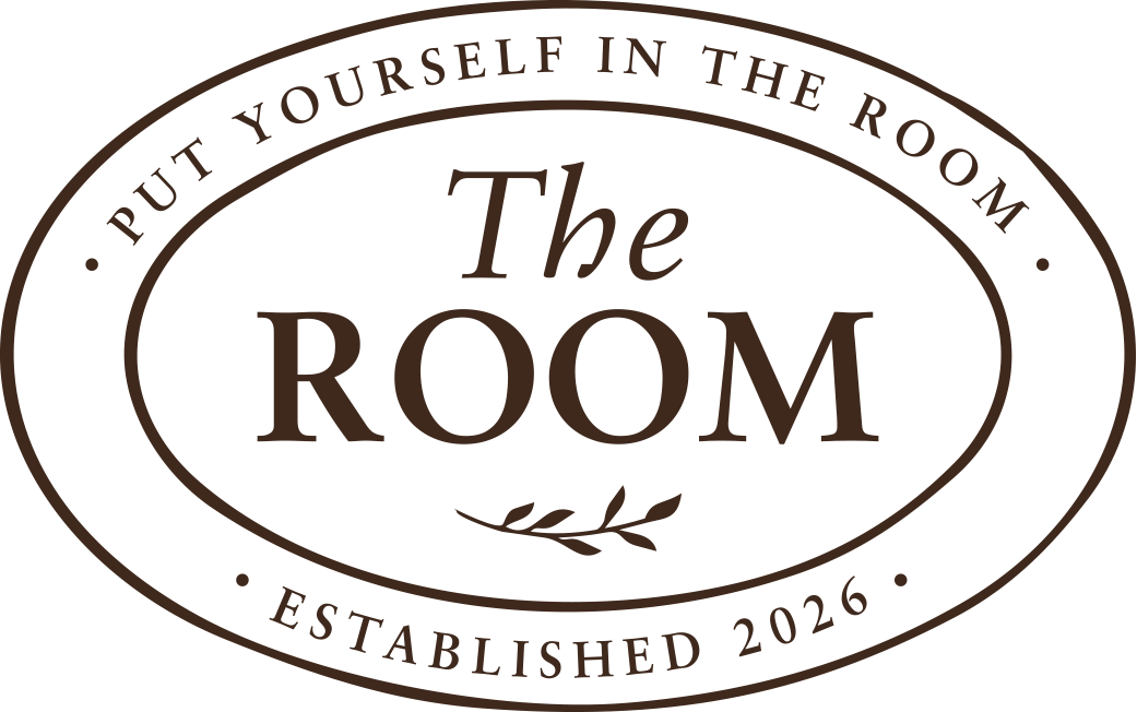 A logo for 'The Room,' featuring the text 'Put Yourself in the Room' and 'Established 2026,' with a decorative leaf element inside a brown and black oval design.