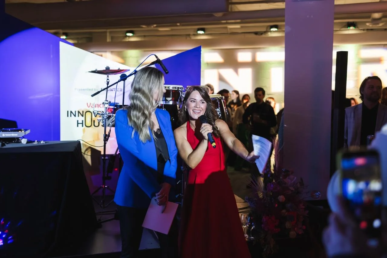 Two women speaking at an event with one using a microphone, one wearing a red dress and the other in a blue blazer, with a drum set and a large screen in the background, and several people in the audience.