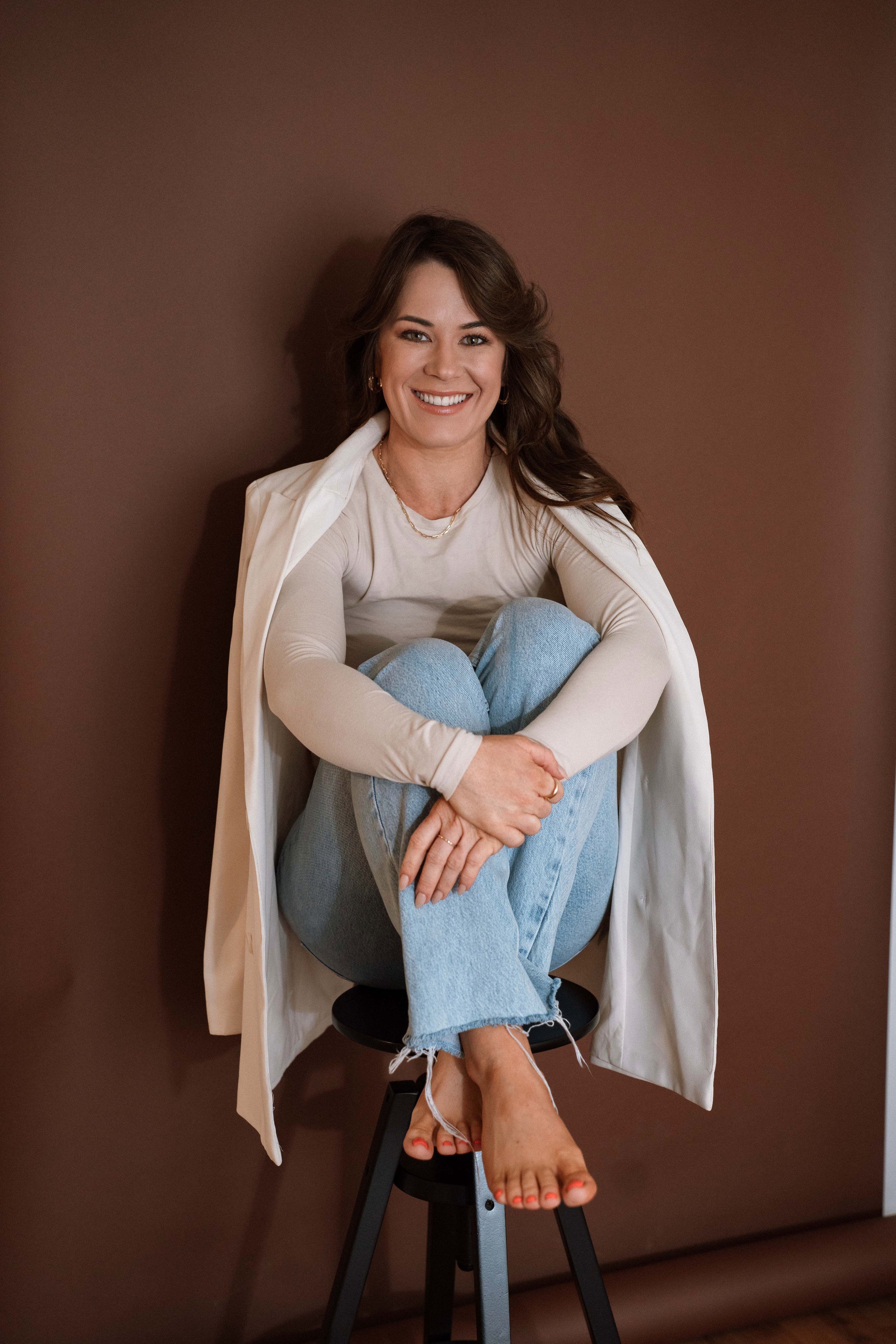 A woman smiling, sitting on a stool with her legs crossed. She has brown hair, is wearing a beige long-sleeved top, blue jeans, and a light-colored cardigan draped over her shoulders. The background is a solid brown wall.