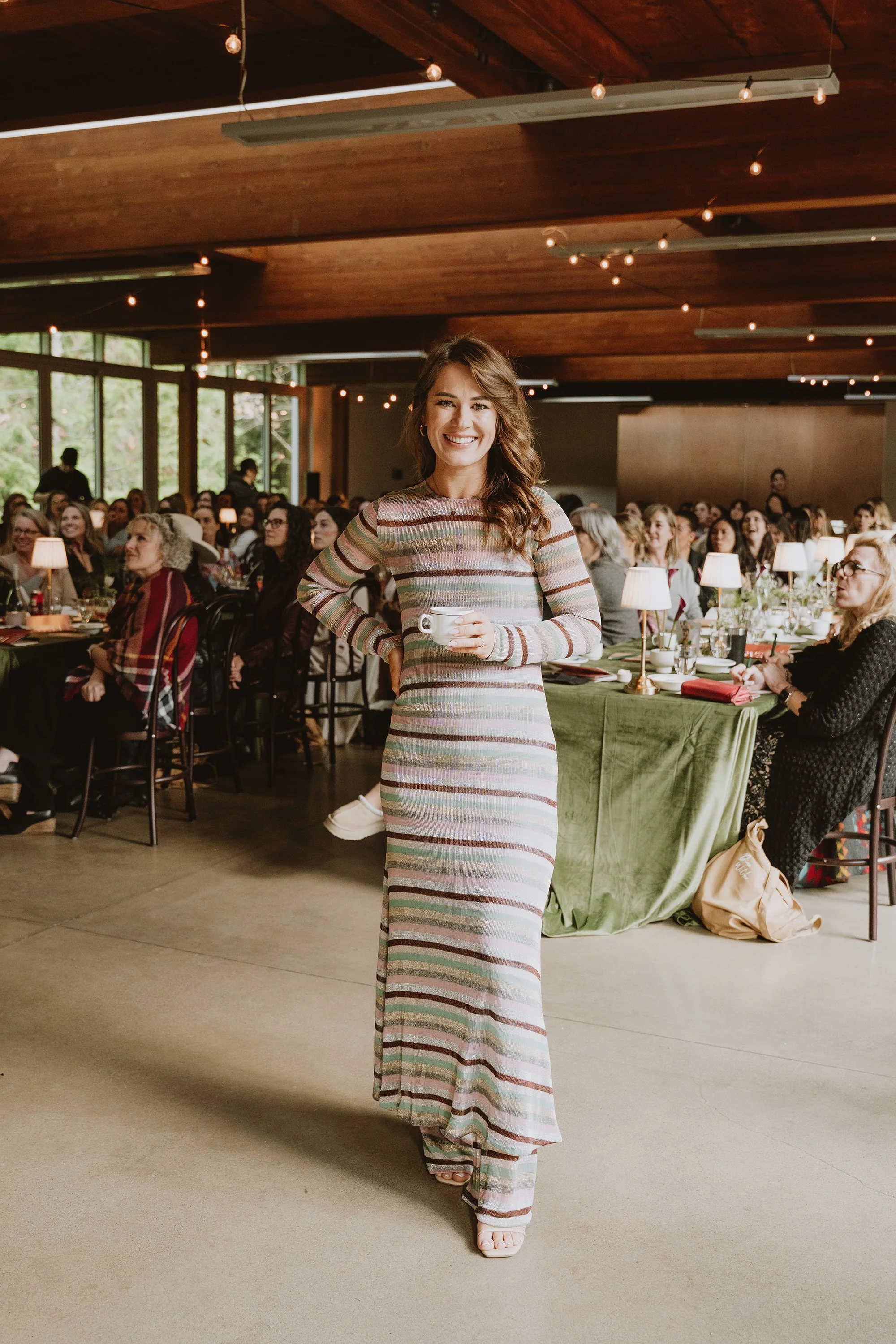 A woman wearing a long, striped dress is smiling while holding a cup, standing in front of a seated crowd at a gathering or event in an indoor venue with wooden ceiling and string lights.