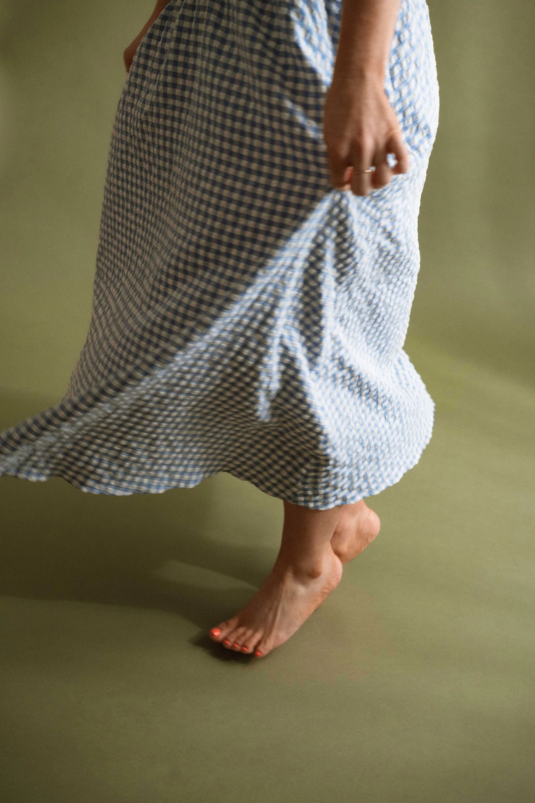 A person with one leg and foot touching the ground and the other leg bent at the knee, wearing a blue and white checkered dress, standing on a green surface against a green background.