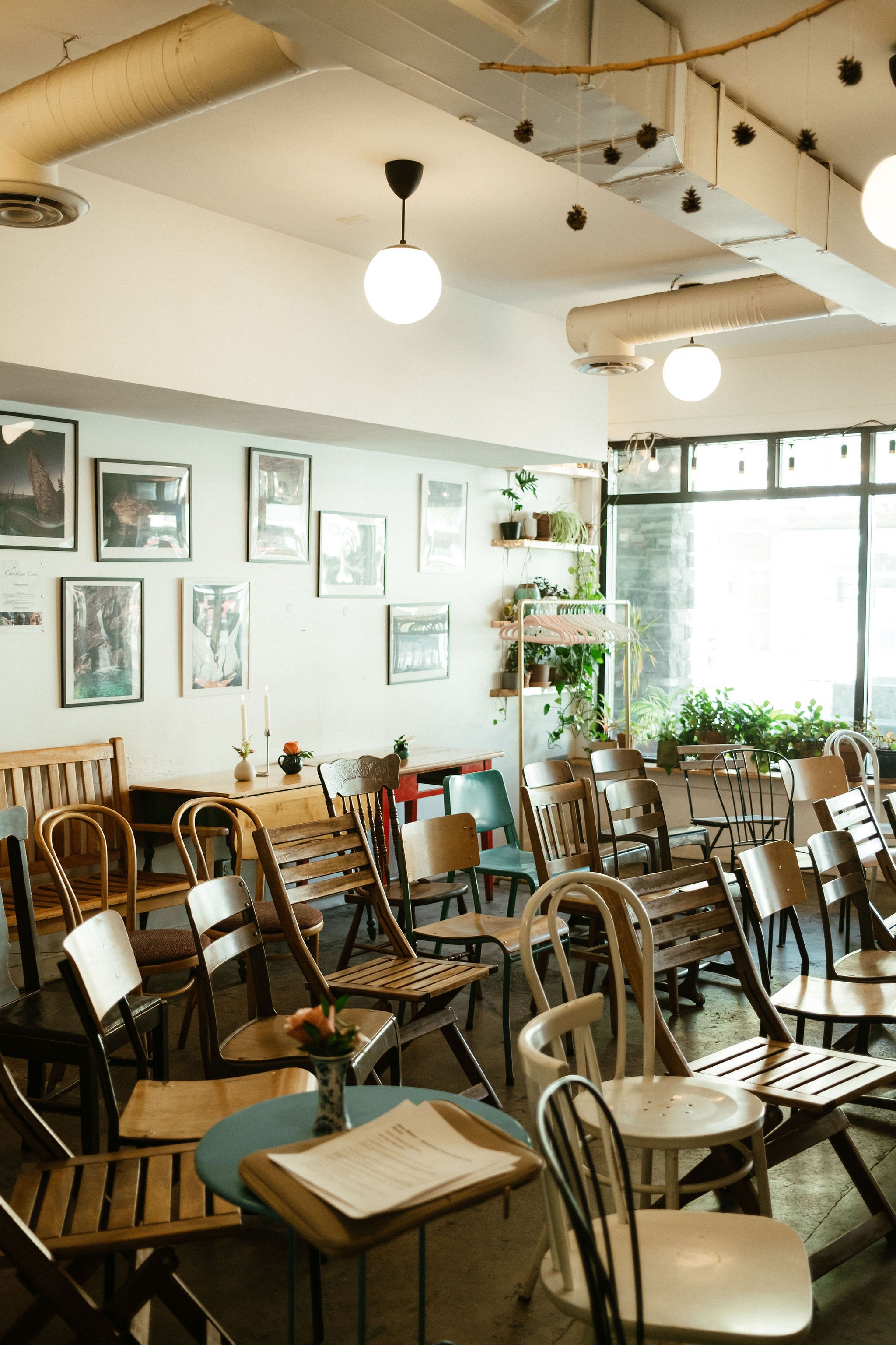 Interior of a cozy cafe or restaurant with various wooden chairs and tables, some with place settings and small flower vases, bright natural light coming through large windows, and framed artwork on the wall, creating a warm and inviting atmosphere.