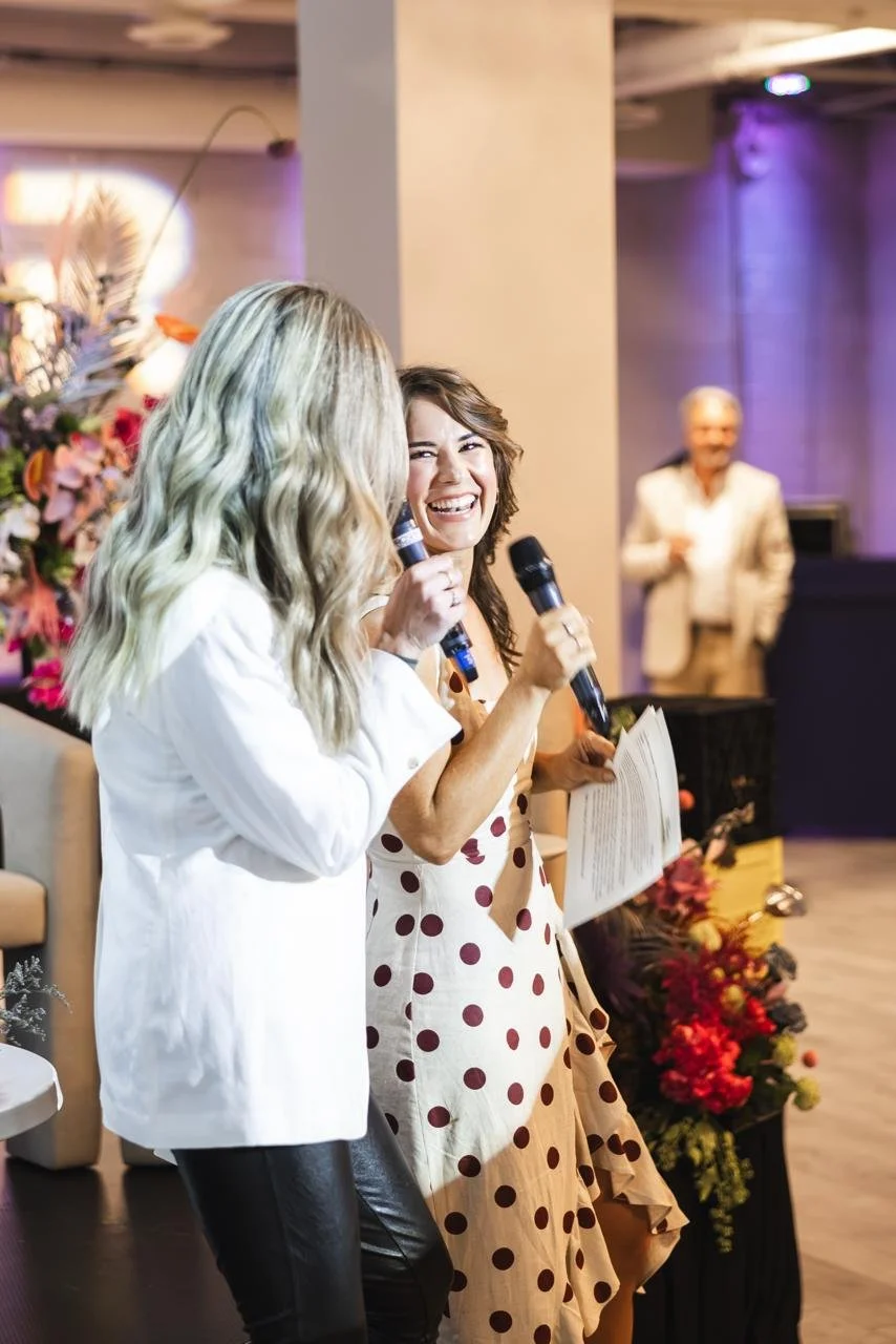Two women smiling and talking with microphones at an indoor event, with one holding a paper, in front of a flower arrangement.