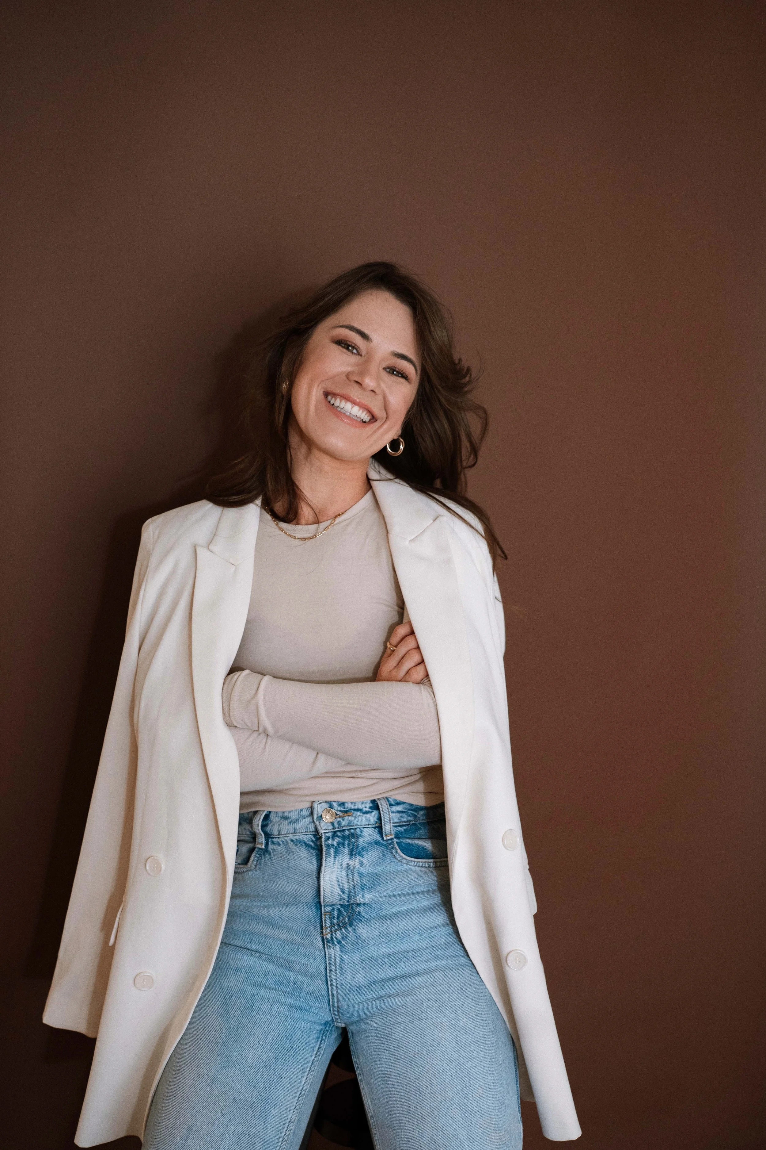 A woman smiling and wearing a white blazer over a beige top and blue jeans, standing with her arms crossed against a brown background.