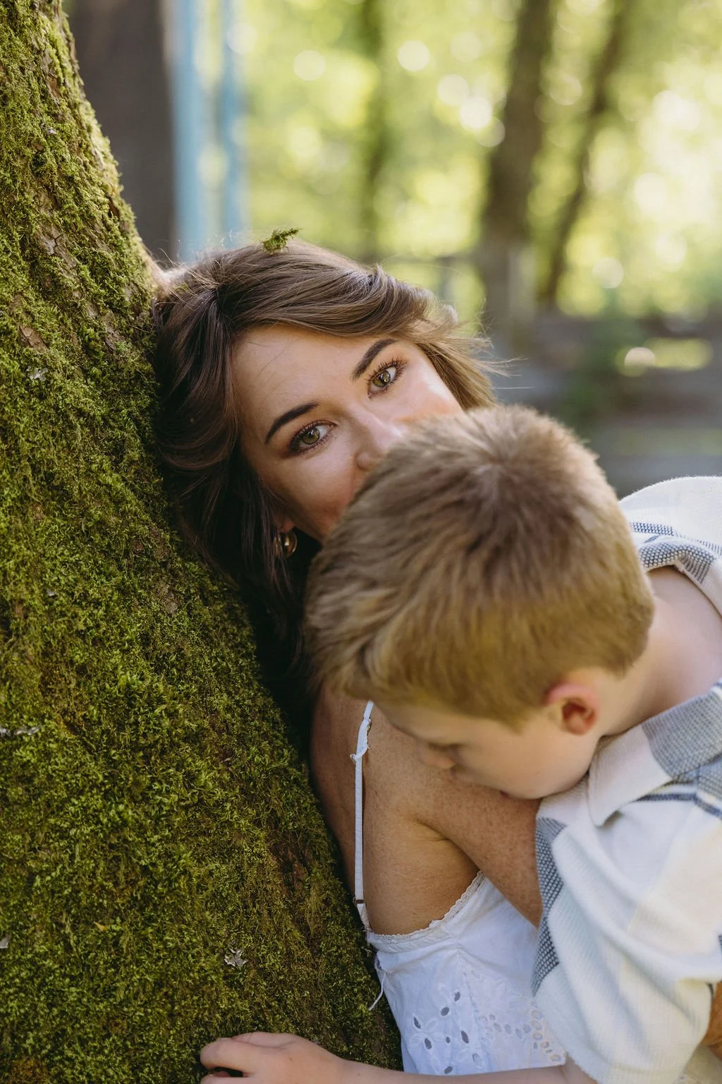 A woman with brunette hair and green eyes lying against a moss-covered tree trunk, looking at the camera while a young boy with short blond hair leans on her shoulder in a forest setting.
