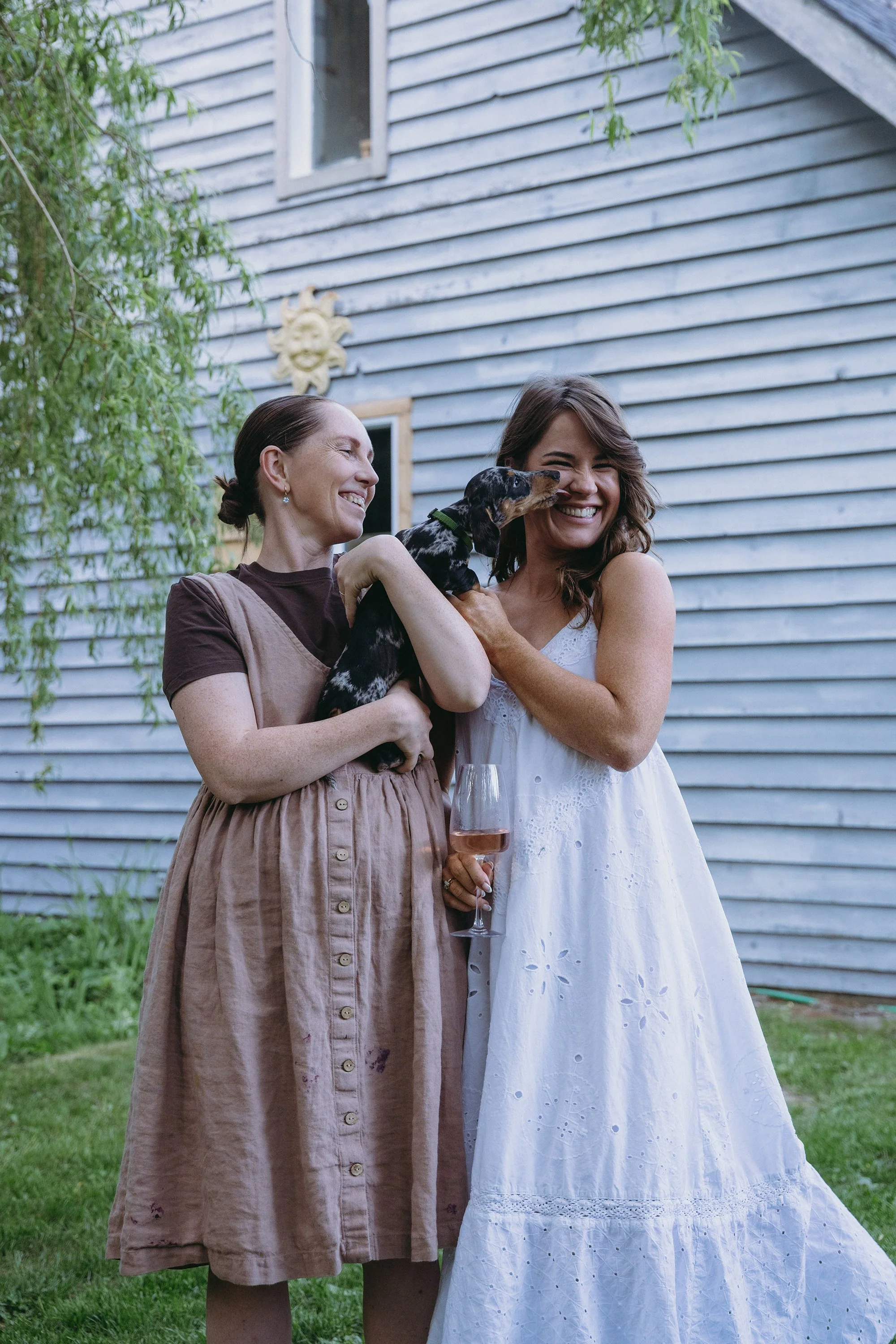 Two women smiling and holding a black and tan puppy. One woman is wearing a brown dress with buttons, and the other a white dress and holding a glass of rosé wine. They are standing outdoors in front of a blue wooden house with a sun decoration on the wall and some green foliage.