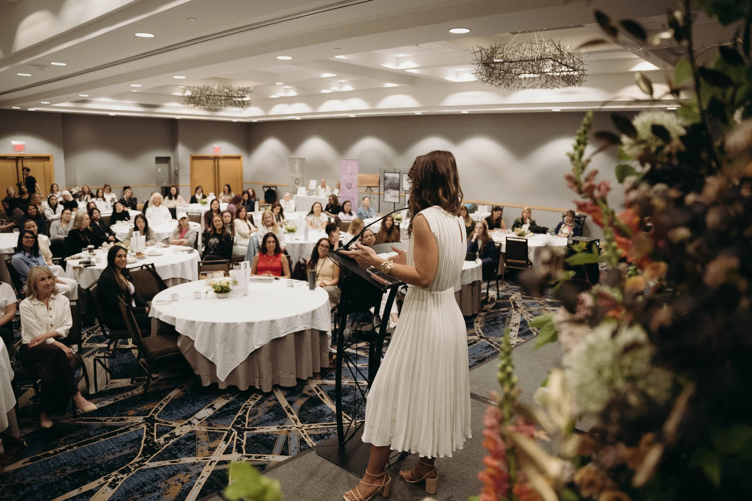 A woman with long, wavy hair in a white dress is speaking at a podium during an event, with a large audience of women seated at round tables in a conference room.