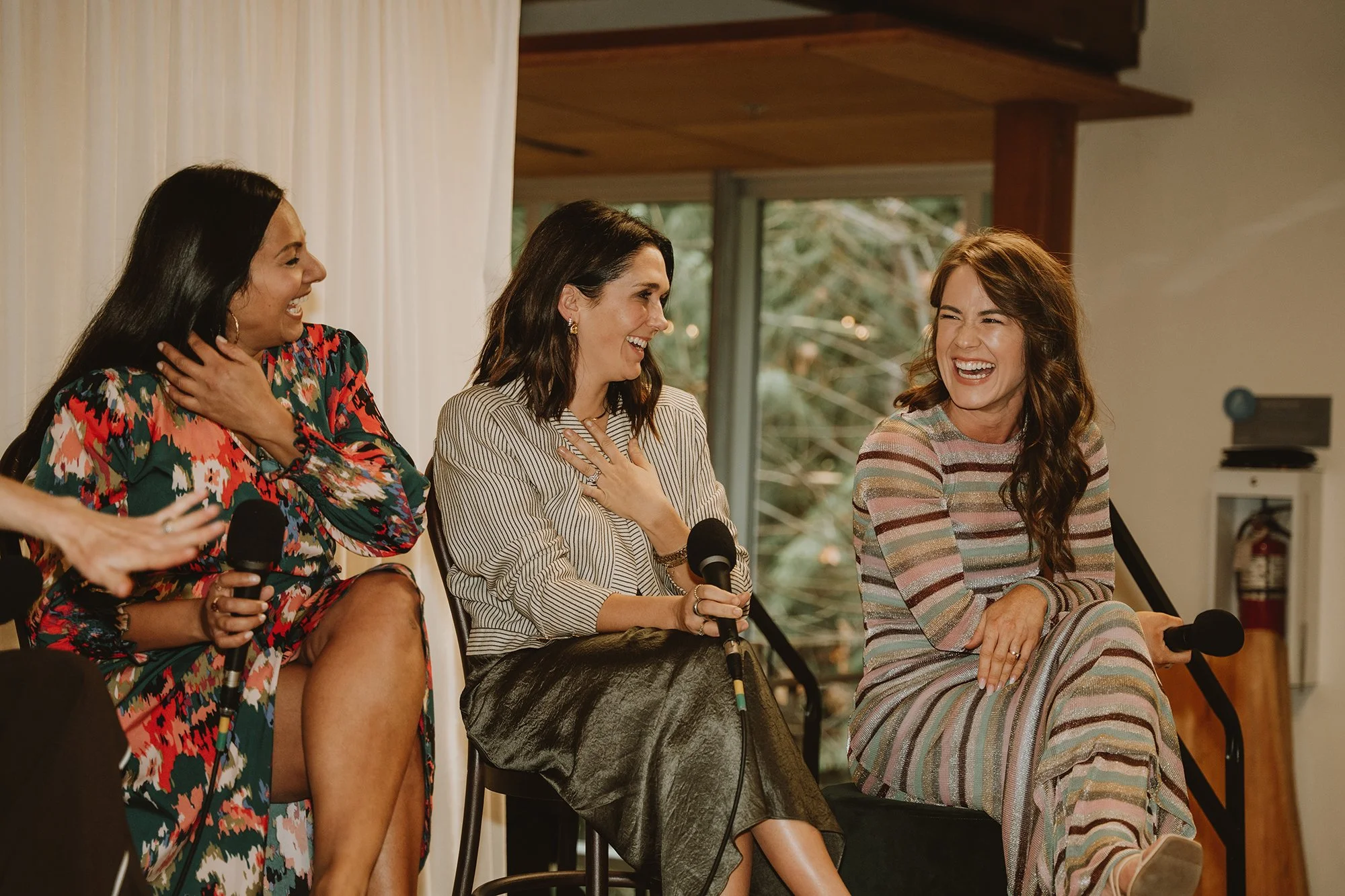 Three women sitting on chairs, laughing and holding microphones, during a panel discussion in a well-lit room with large windows and greenery outside.
