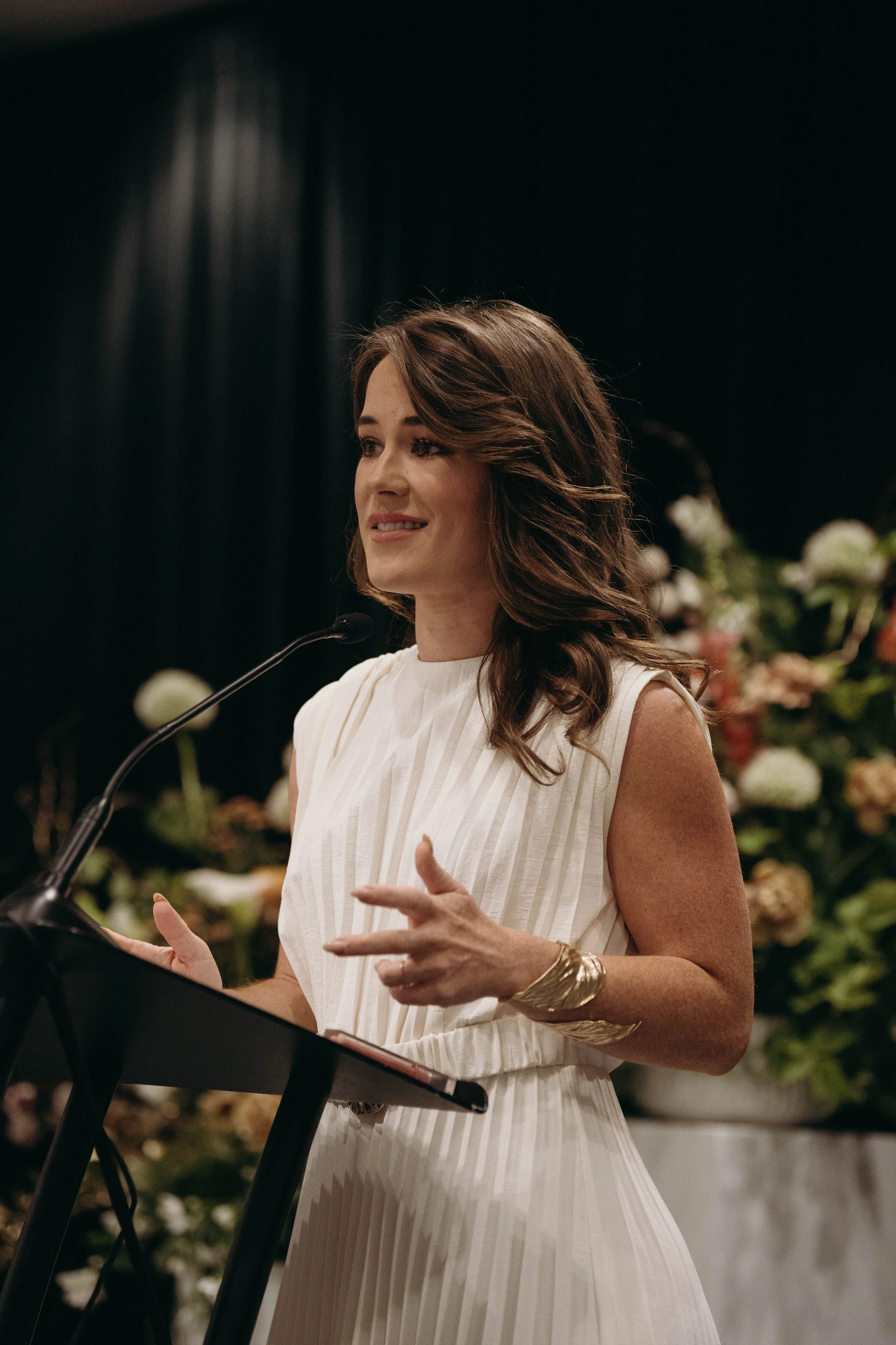 A woman with brown wavy hair speaking into a microphone, wearing a white sleeveless dress and a gold bracelet, standing at a keyboard, with a backdrop of flowers in a dark room.