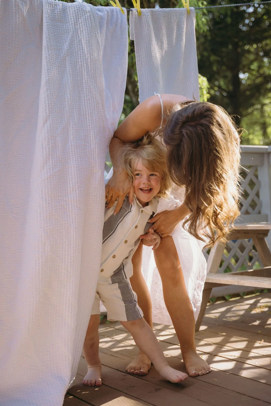 A woman and a young boy are playing hide and seek behind white curtains outdoors, with smiles and a tree in the background.