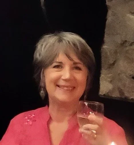 A woman with short gray hair smiling and holding a glass of wine or champagne in a dimly lit setting.