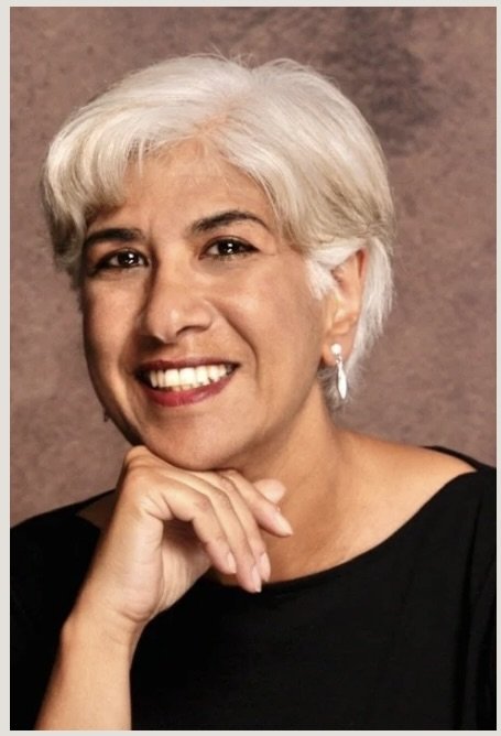 Portrait of an older woman with short white hair smiling and resting her chin on her hand, wearing a black top and silver earrings.