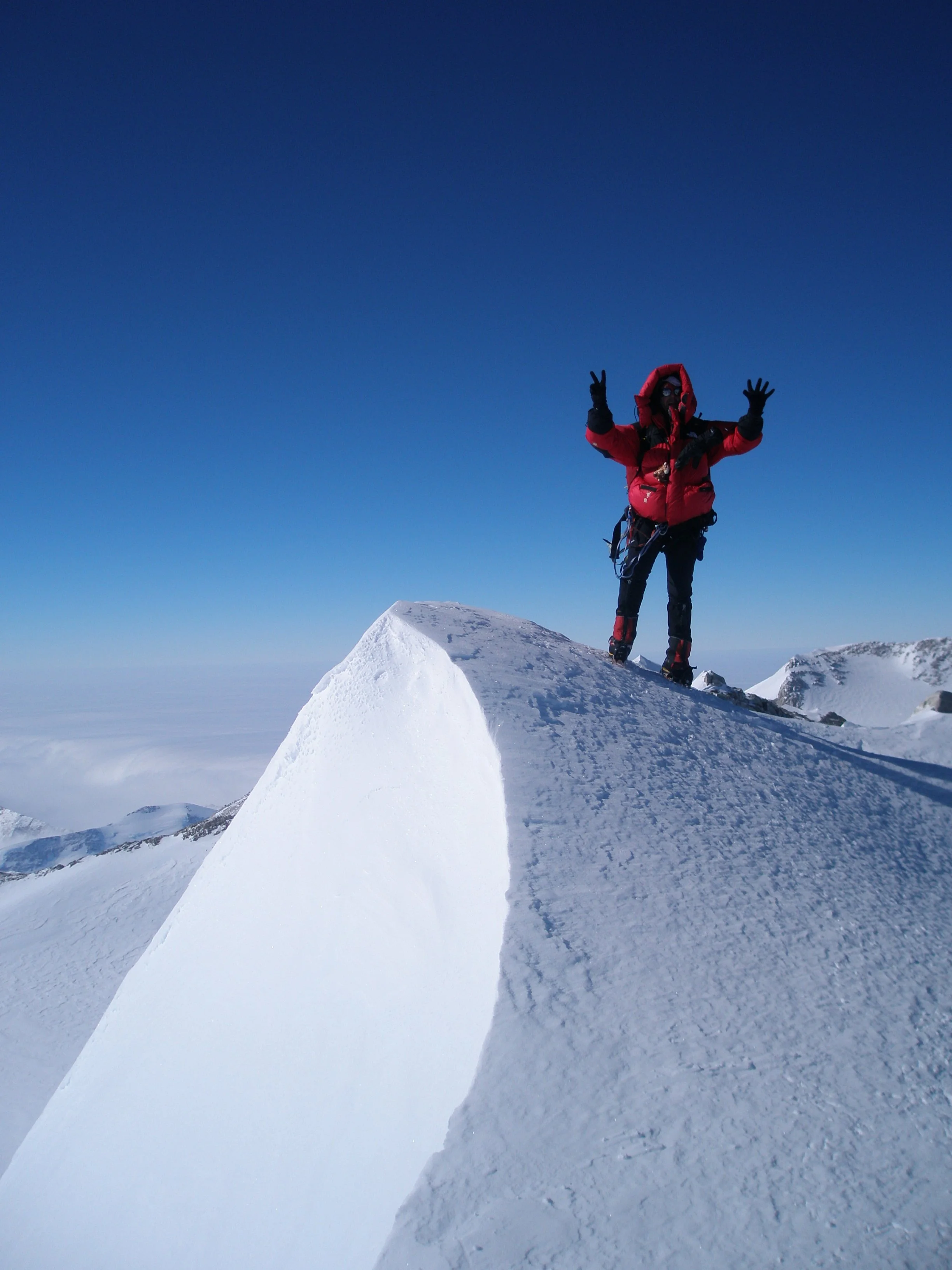 A person dressed in climbing gear, including a red jacket and black gloves, stands on a snowy mountain summit, making a peace sign with both hands against a clear blue sky.