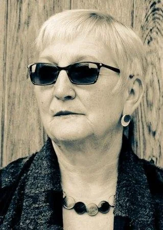 A woman with short blonde hair wearing dark sunglasses, a dark textured jacket, and a necklace with large black beads, standing against a wooden background.